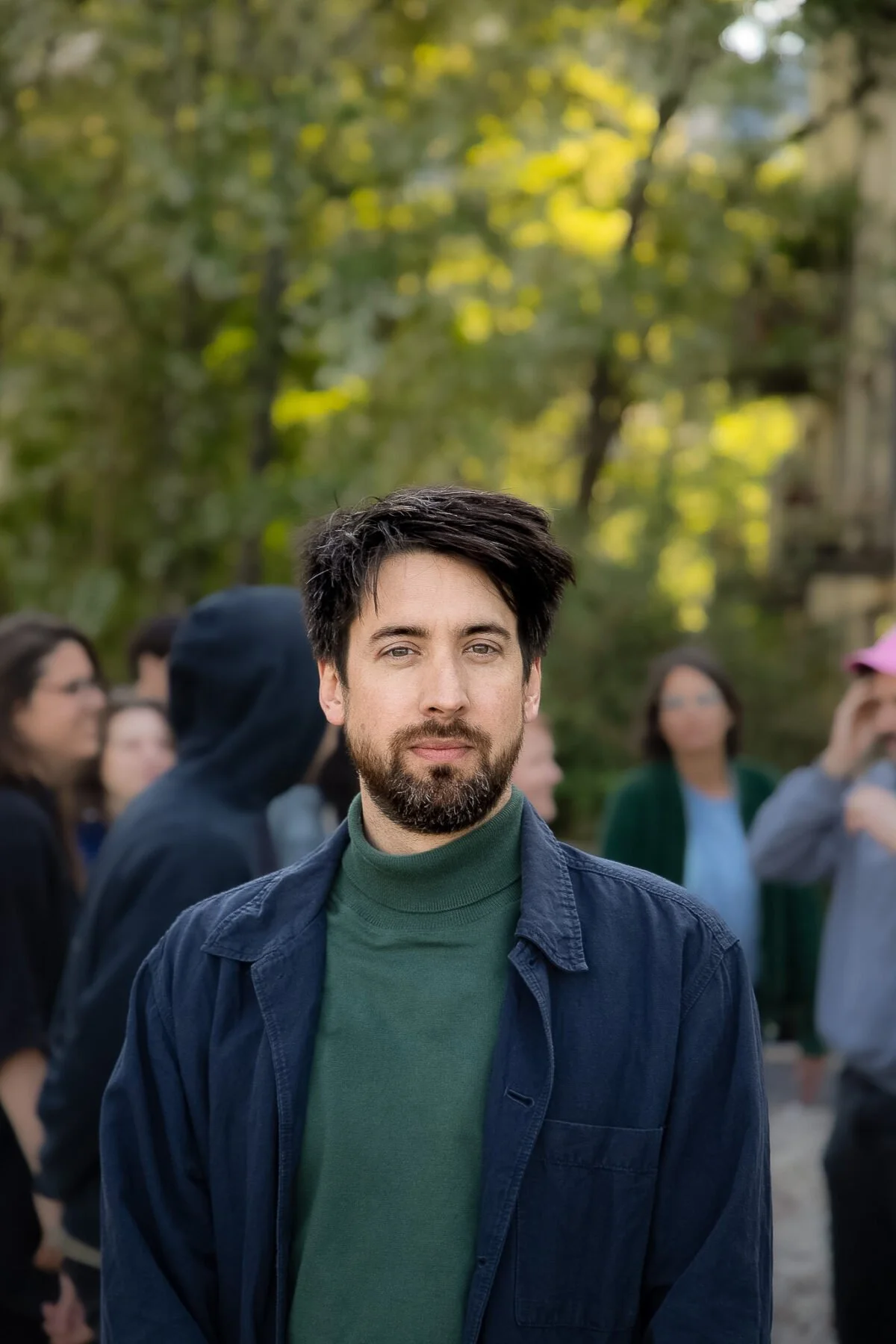 Ein junger Mann mit dunklem Haar und Bart steht im Vordergrund, im Hintergrund versammelte Menschen in einer grünen Parkumgebung.