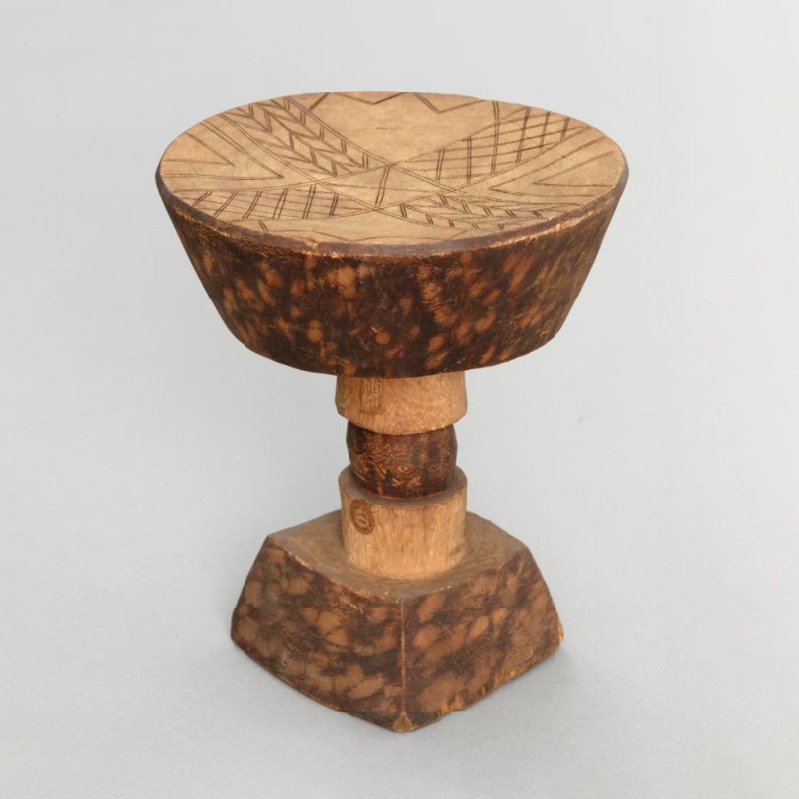 A stool, cut from a solid block of light wood. It has a square base with a central pillar to support the top, which is an inverted truncated cone.