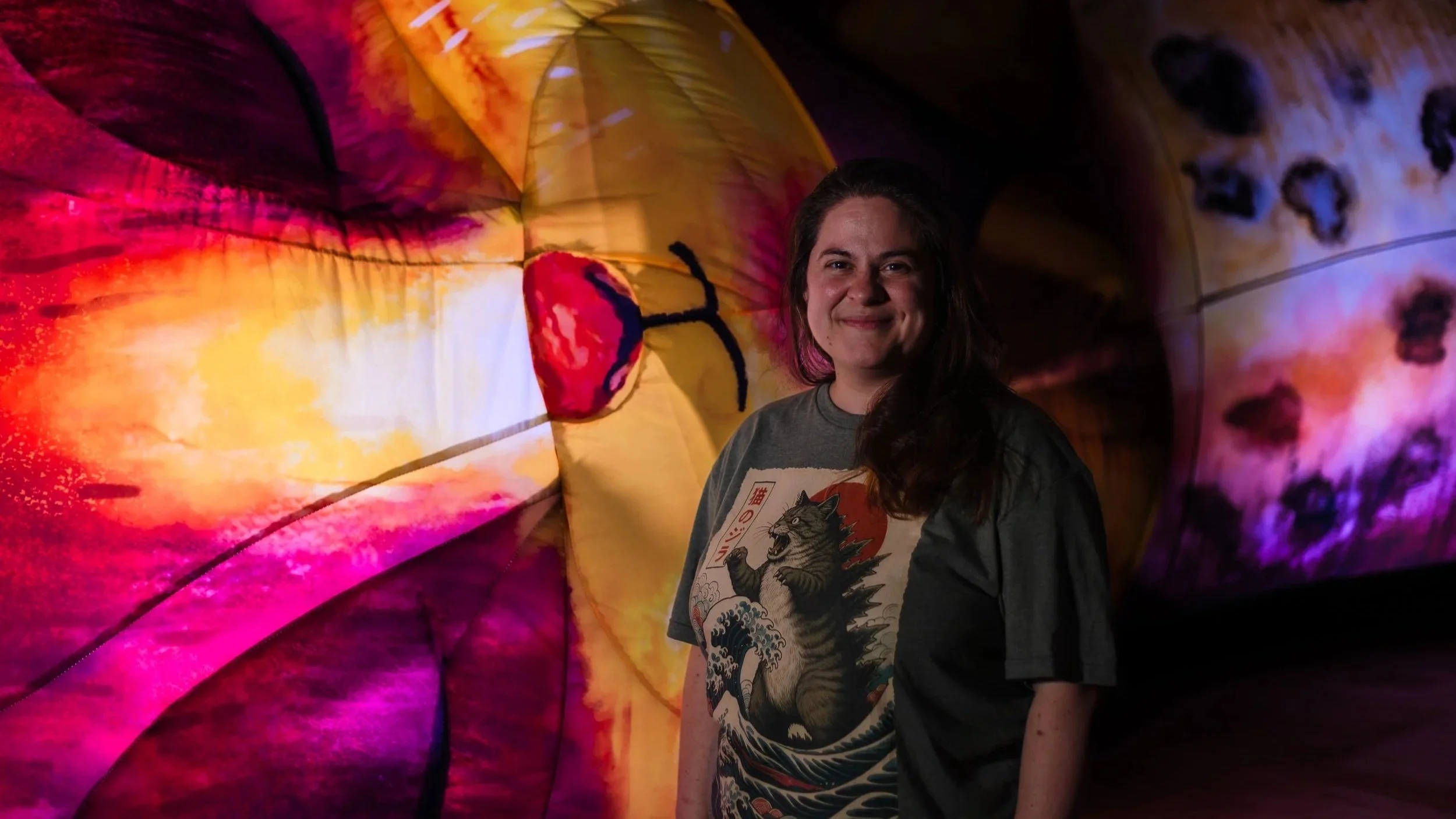 A woman with long dark hair smiling, standing in front of colourful, illuminated, inflatable cat.