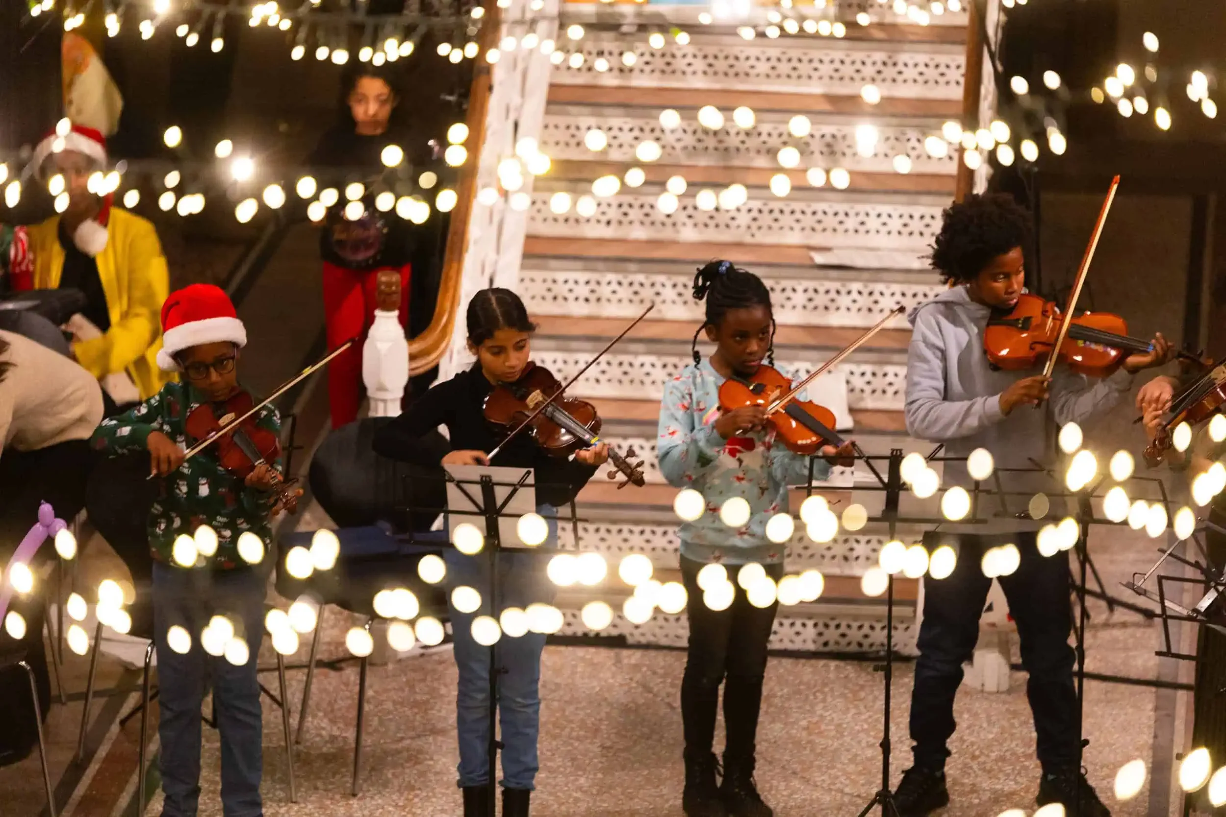 Children playing violins during Olympias Music Foundation's winter concert in Maanchester Museum's Living Worlds gallery, with christmas lights and a staircase in the background.