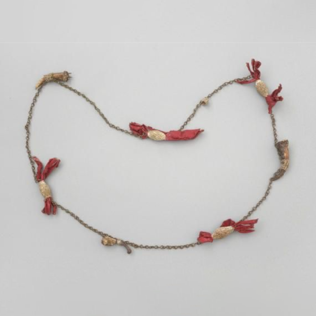 A Necklace made from a brass chain with attachments of red cloth and jawbones attached at intervals by bands of white buttonholing.