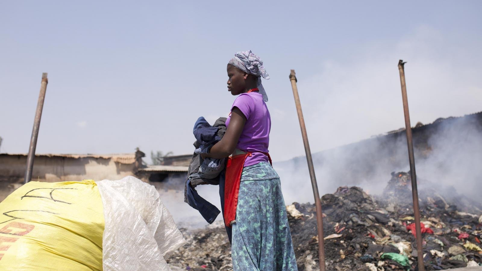 A person holding clothes walks past huge mounds of smouldering fabric waste.