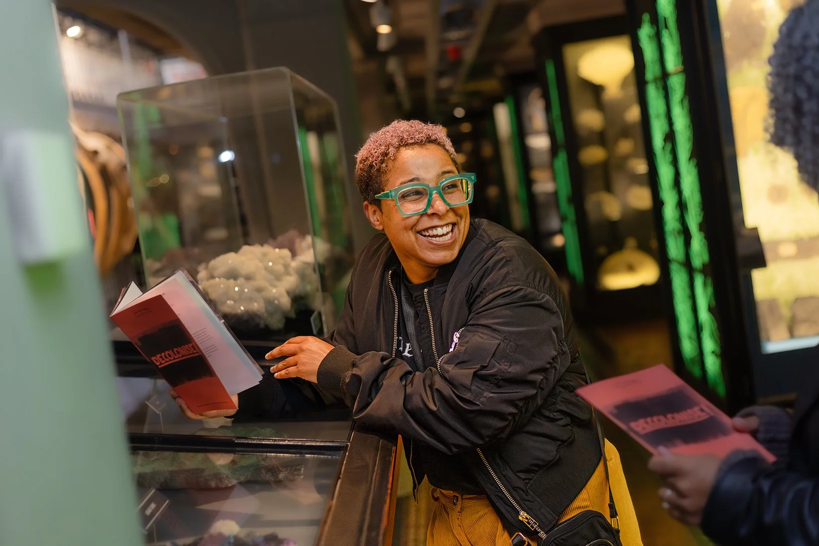 A person smiling while holding one of Manchester Museum's Decolonise! trail leaflets, leaning on cases in the Nature's Library gallery.