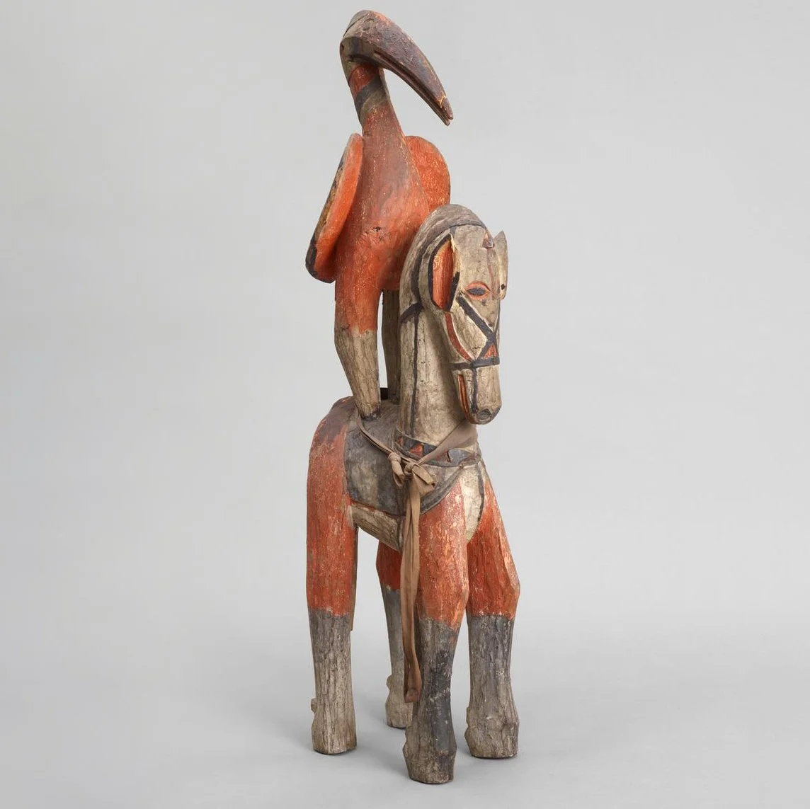 A wooden carving of a long-legged horse with an ibis mounted on it's back. The horse is painted black, orange and white, with it's harness picked out in black. The ibis is painted in the same colours.