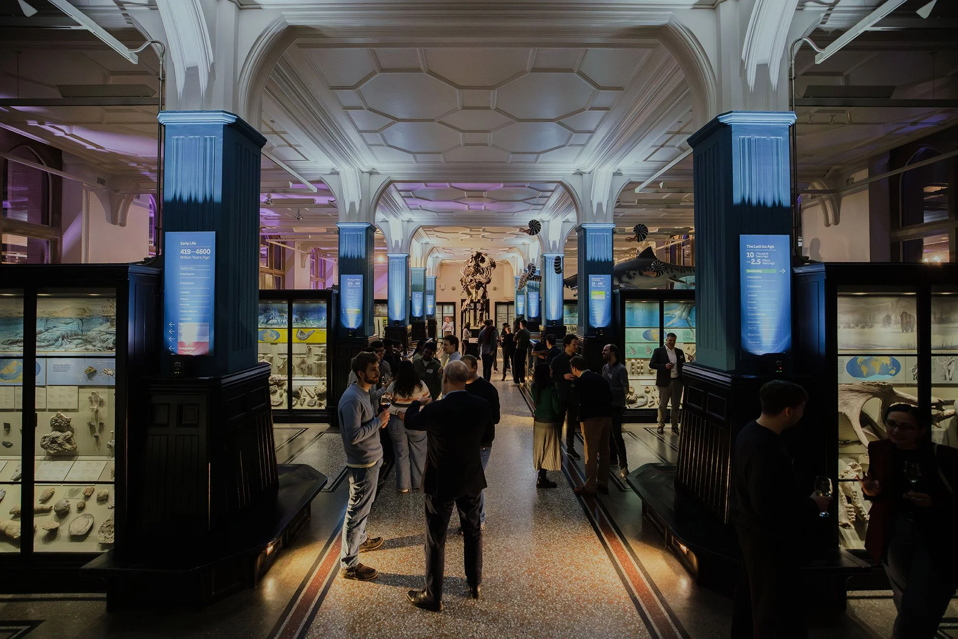 People enjoy themselves at a drinks reception in the Fossils and Dinosaurs gallery at Manchester Museum. The room is lit in blue and purple and Stan the Tyrannosaurus rex can be seen in the background.