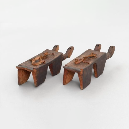 A wooden artefact, possibly a ceremonial sandal. Cut from a single block of wood. Rectangular top with four short, broad, flat legs.
