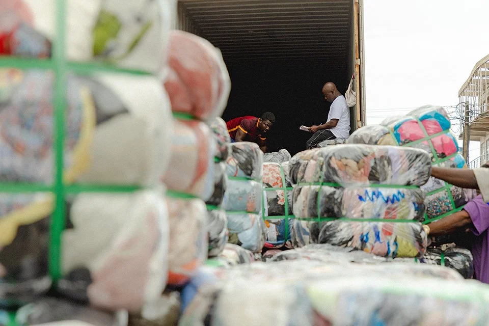 Bales of clothes are stacked in the foreground, with two people sat in a truck in the background.