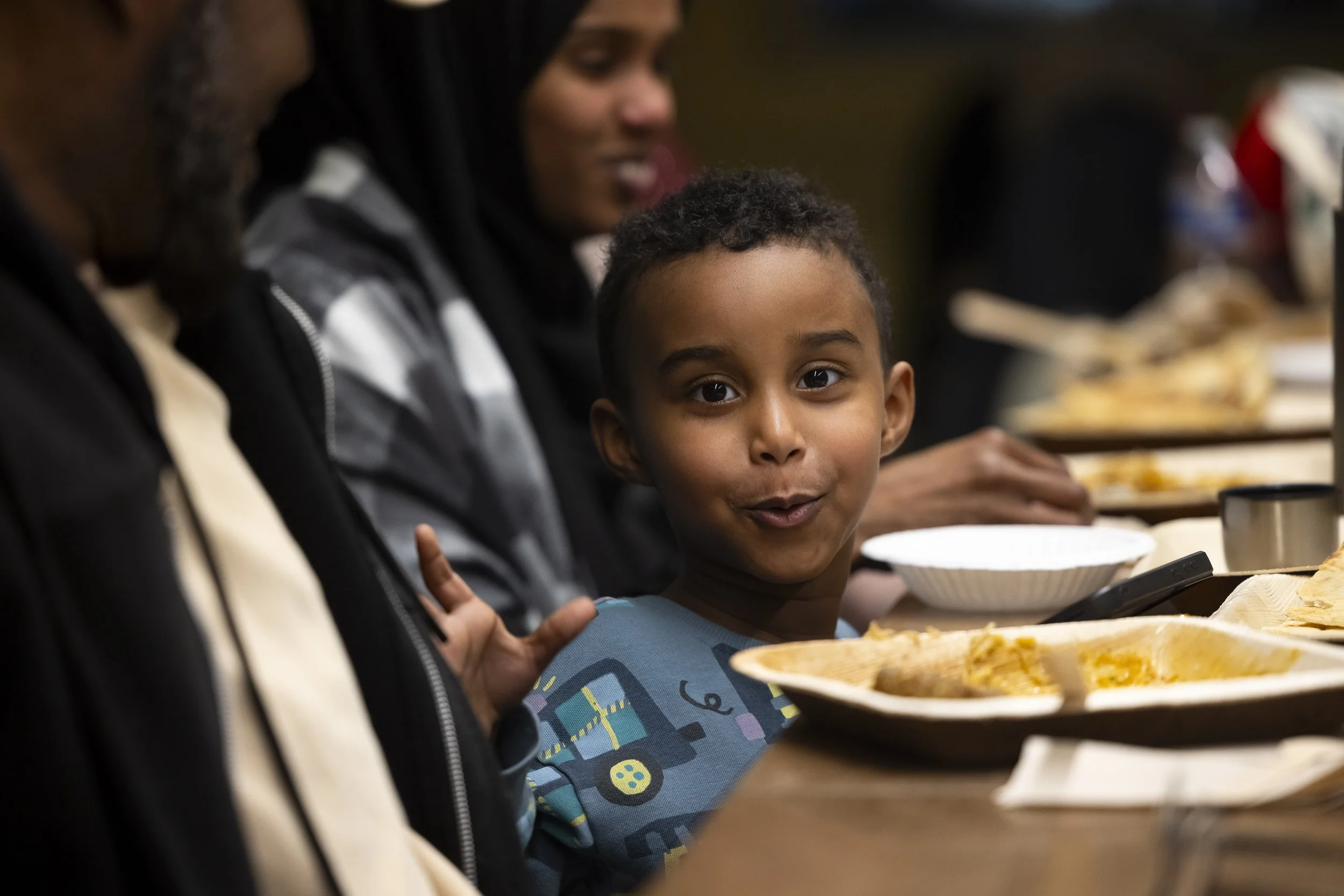 A young child smiles while sat at a table with a plate of food in front of them and adults sat either side.