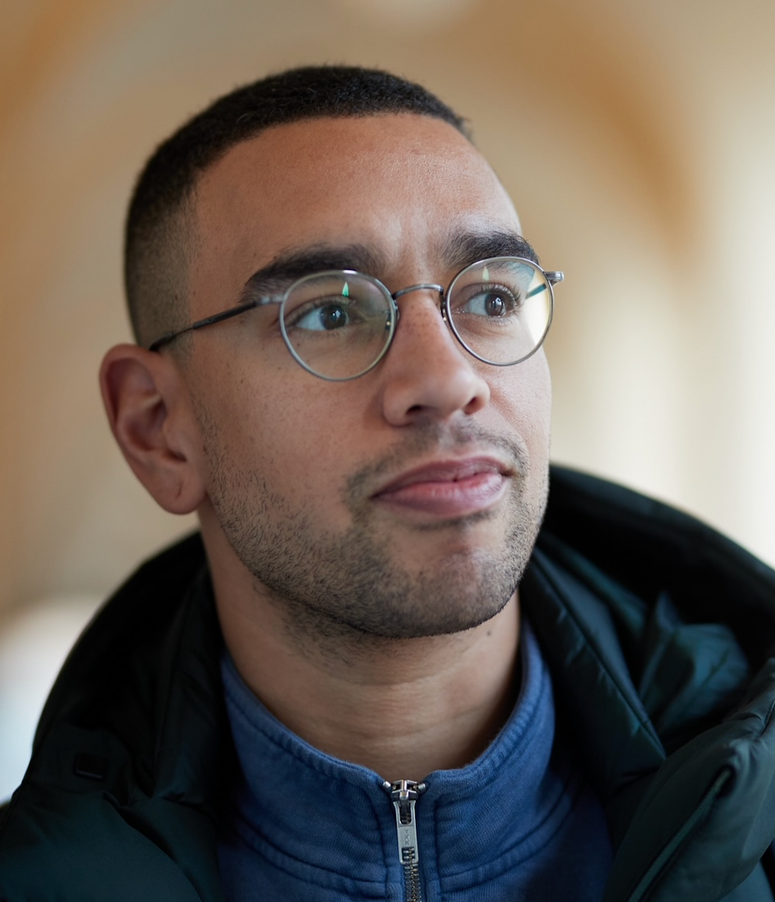 A young man with glasses and a beige shirt standing with arms crossed, smiling in front of a colorful background with flags and text.