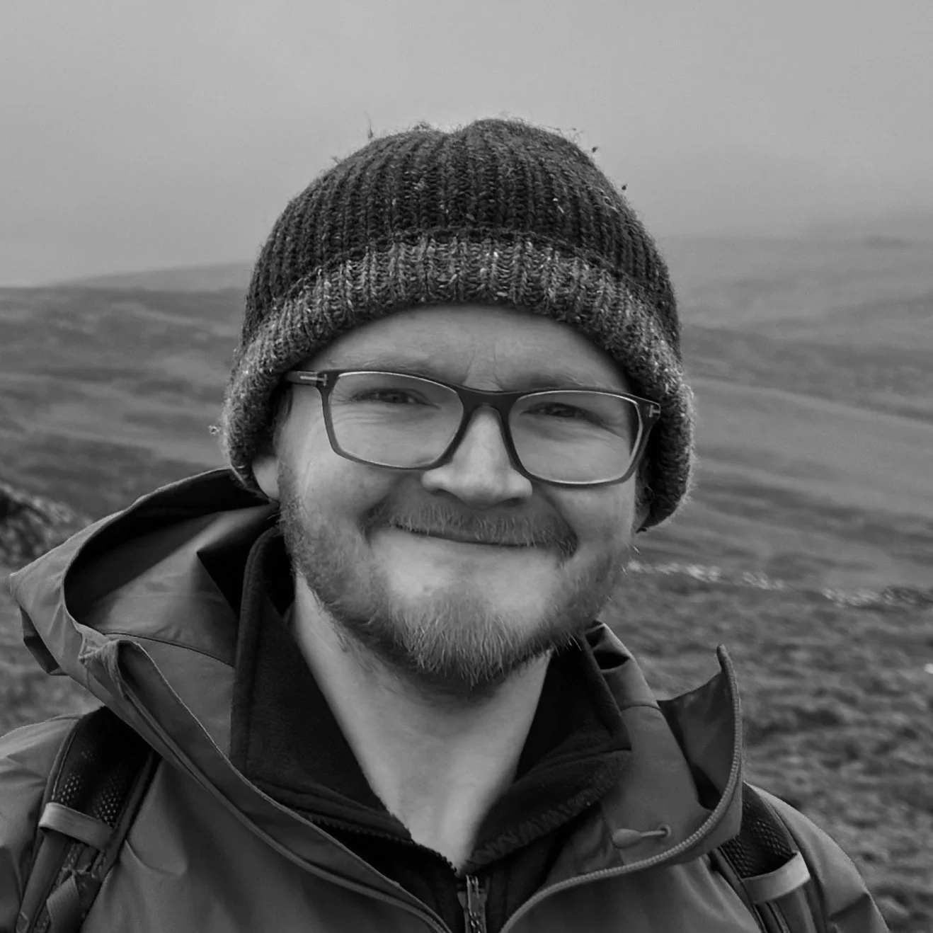 Chris Griffiths, Curator of Archaeology at Manchester Museum, smiling and wearing a beanie outdoors with rolling hills in the background.