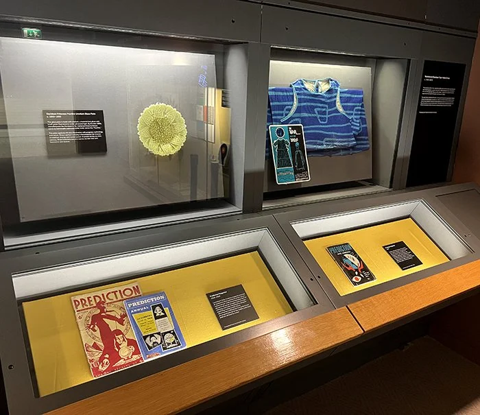 Display case with a yellow background showcasing vintage magazines and informational plaques within the exhibition Artefacts of Prediction in the Research Gallery at Manchester Museum.