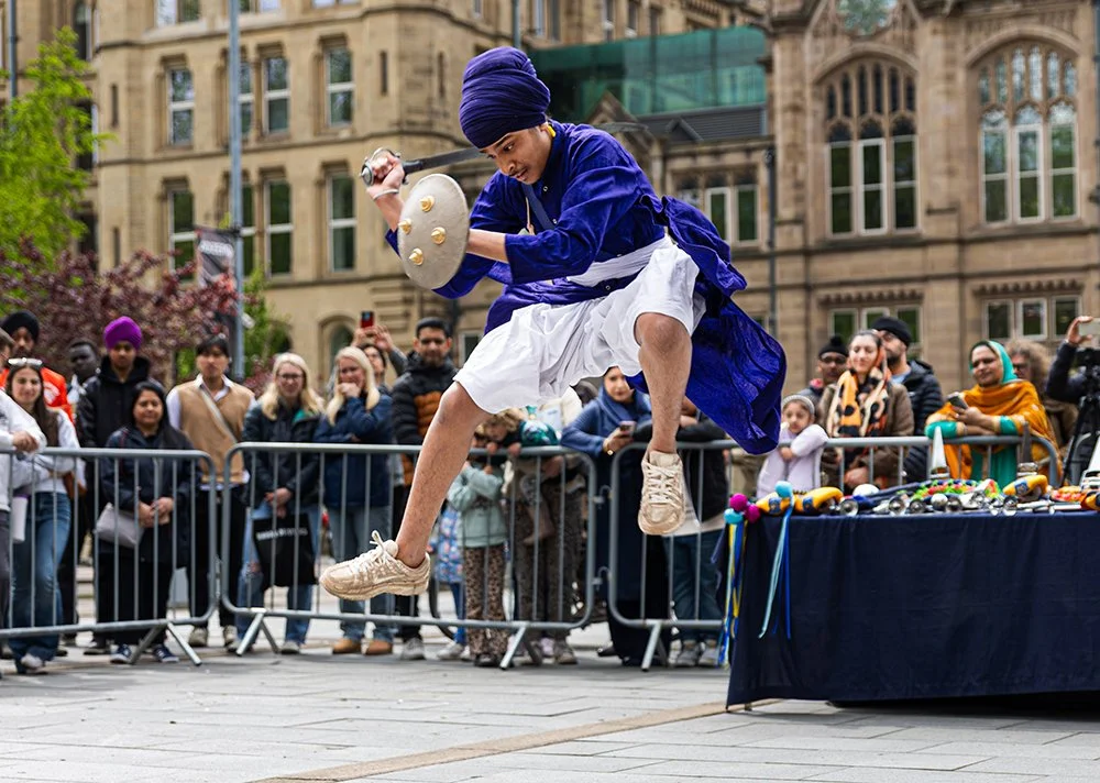 A Gatka martial arts performer wearing traditional Sikh dress in blue and white jumps in the air while holding a sword and shield.