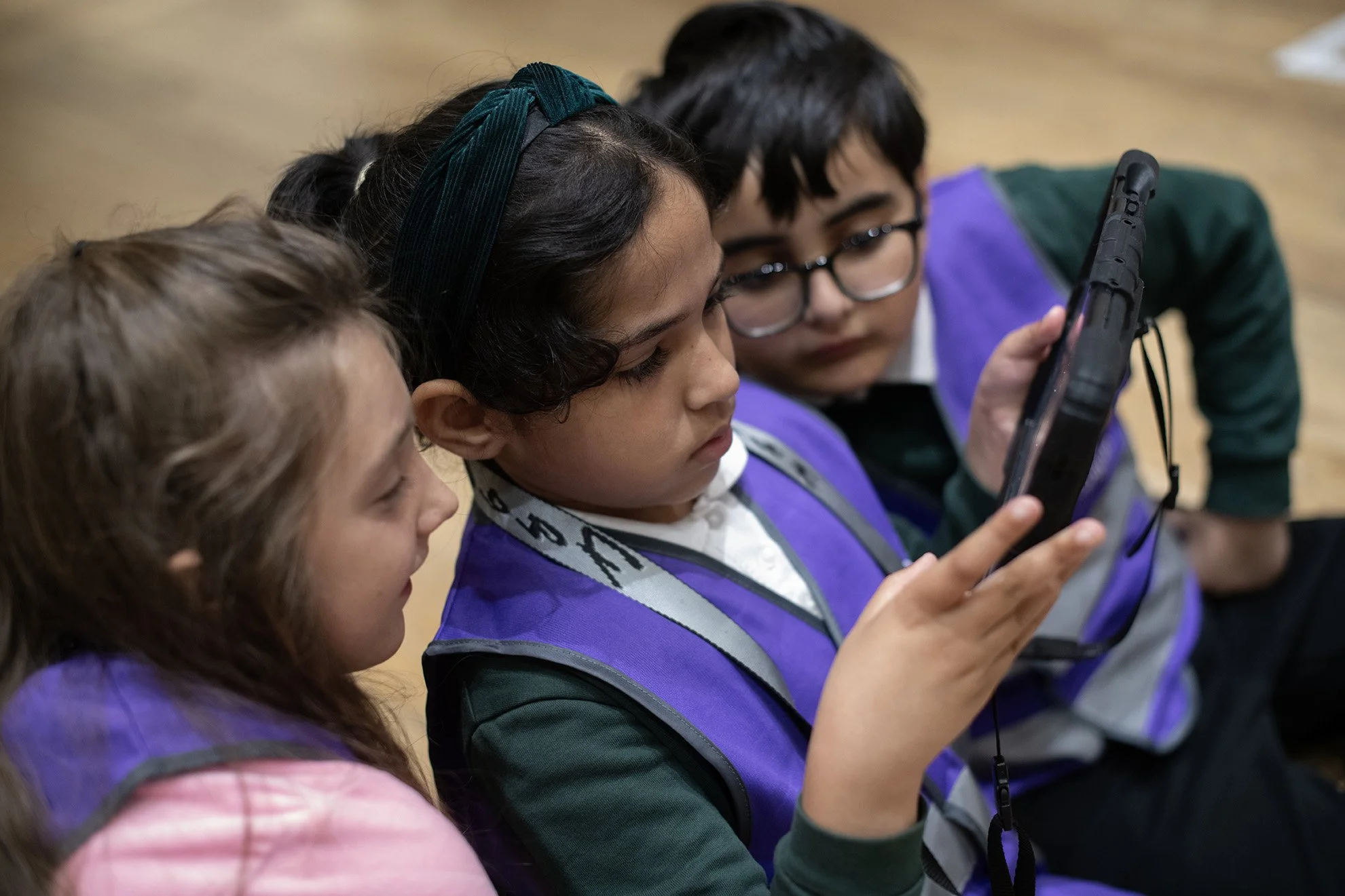 Three children sitting closely together, looking at a tablet device. They are wearing school uniforms and are indoors at a museum.