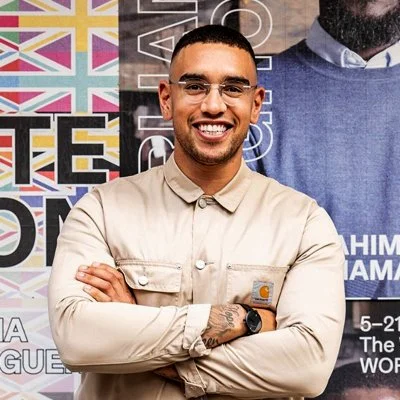 A young man with glasses and a beige shirt standing with arms crossed, smiling in front of a colorful background with flags and text.