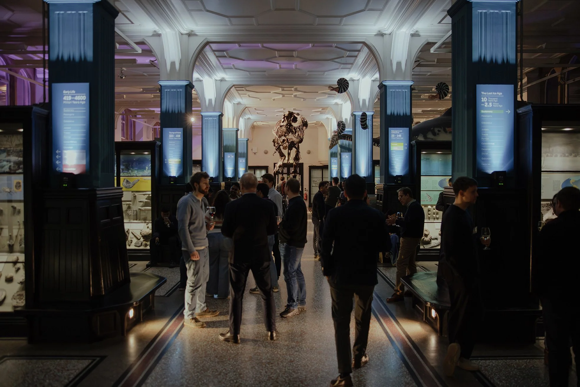 People chat and enjoy themselves at a drinks reception in the atmospheric Fossils and Dinosaurs gallery at Manchester Museum. Stan, the Tyrannosaurus rex, can be seen in the background.