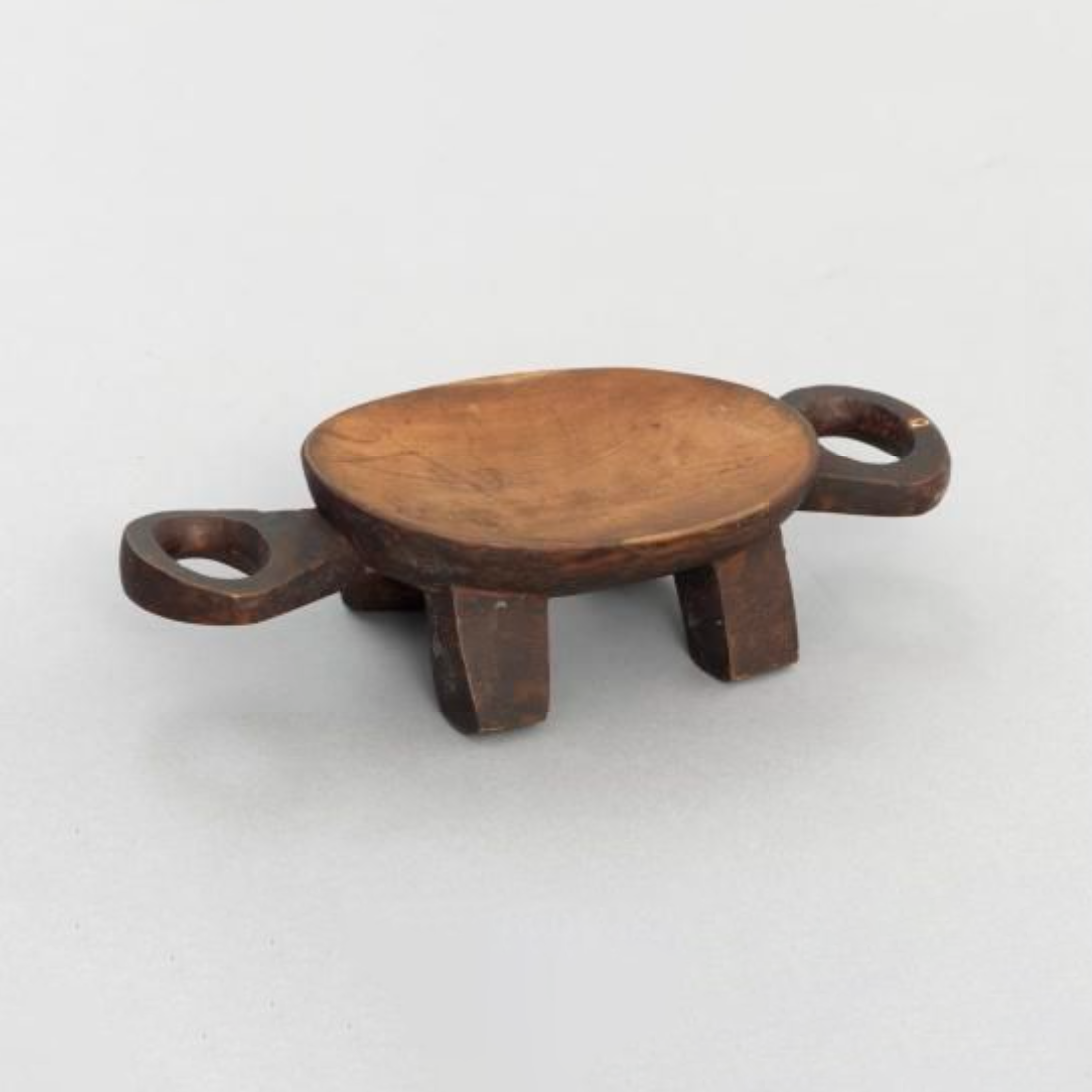 A shallow oval, wooden dish standing on four short, thick legs, with a large, projecting ‘bottle opener’ shaped handle at either end.