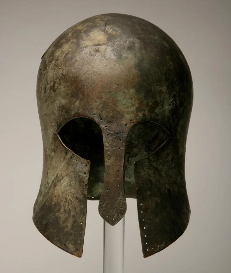 Ancient bronze helmet, possibly Greek or Roman, with a decorative crest and facial protection, displayed on a stand.