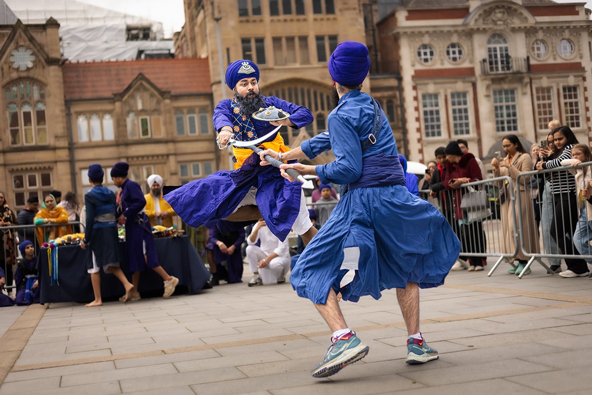 Two people wearing traditional Sikh costumes in bright blue perform a Gatka martial arts demonstration. One jumps in the air holding a sword, while the other holds out a spear.