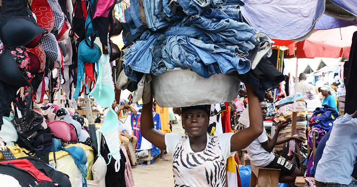 A person walks through a market packed with clothes with stacks of jeans balanced on their head.
