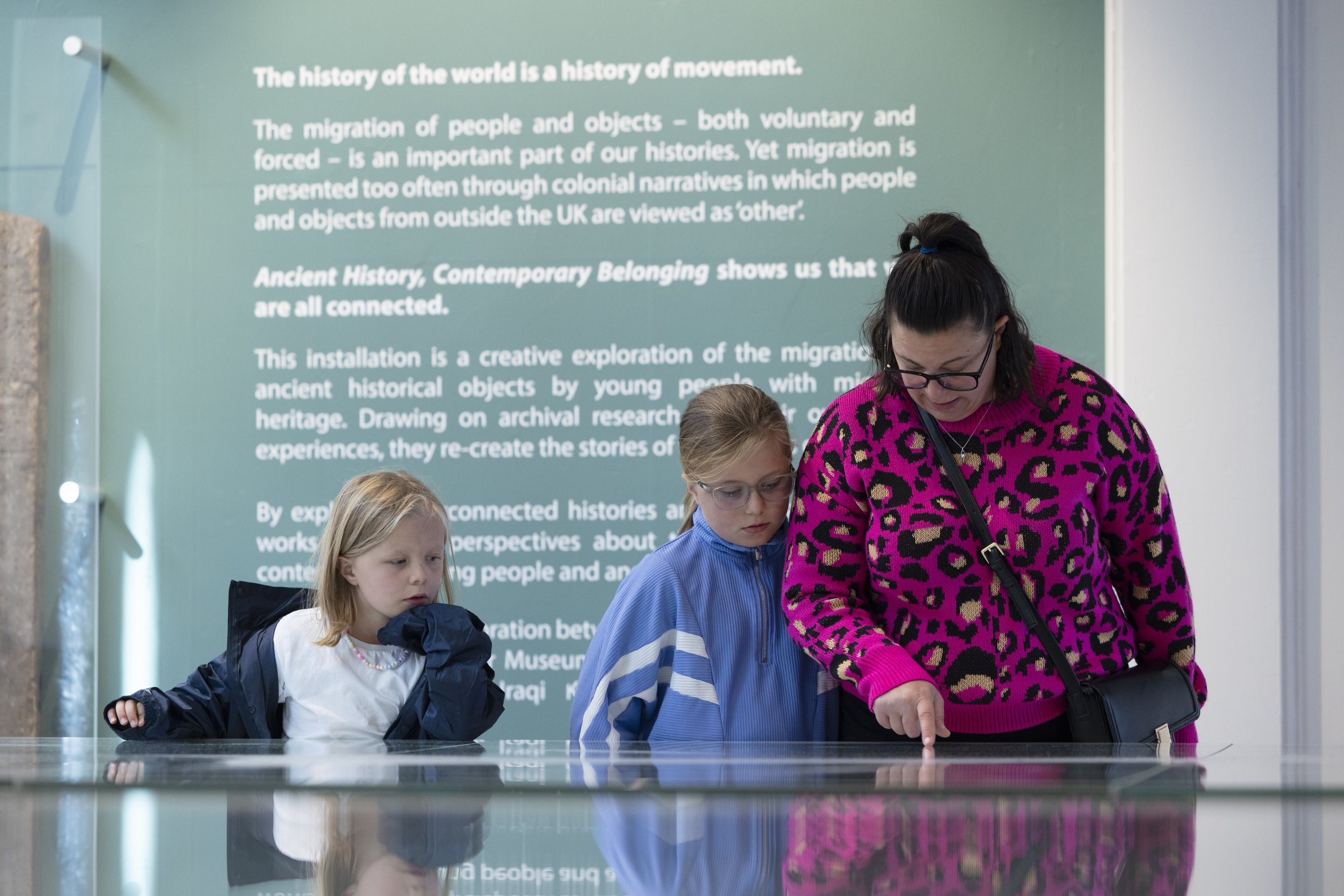An adult visitors points into a glass case in the Archaeology gallery at Manchester Museum while talking to two children.
