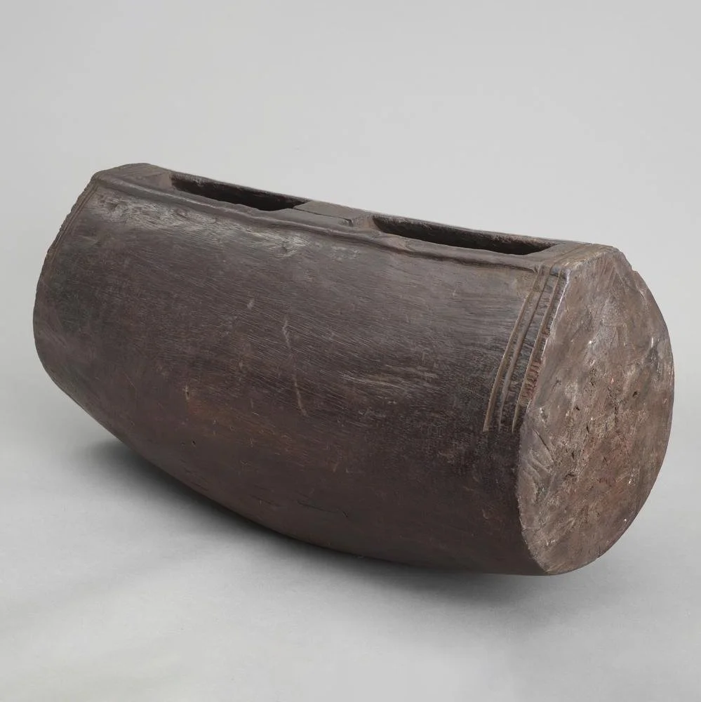 A large slit gong cut from a single block of heavy, hard wood, probably a section of tree trunk. It is barrel shaped and the interior is hollowed out and there is a slit left in one side with a short handle left in the centre.