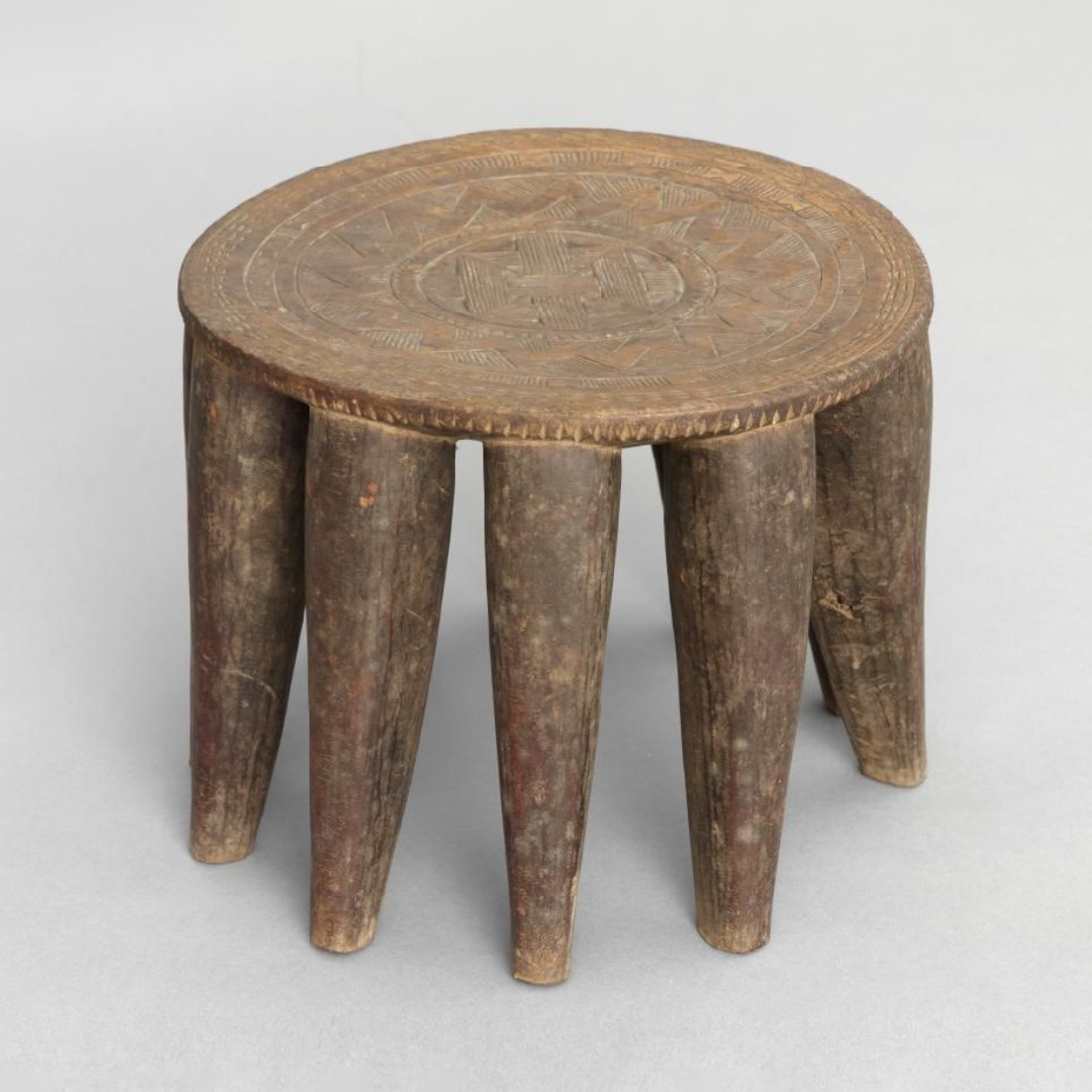 A circular stool with ten round, tapering legs, all cut from one solid block. The top is covered with shallow carving.