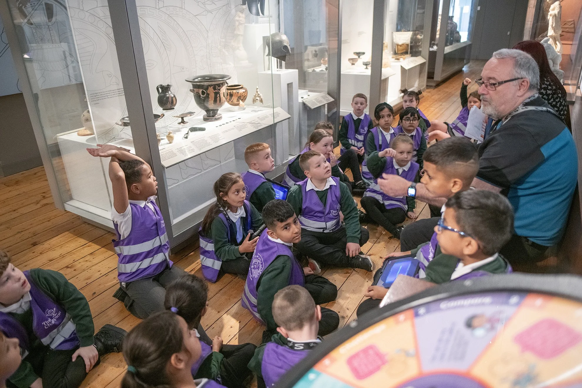 Group of school children in purple vests sitting on wooden floor listening to a tour guide in a museum exhibit room with glass display cases.