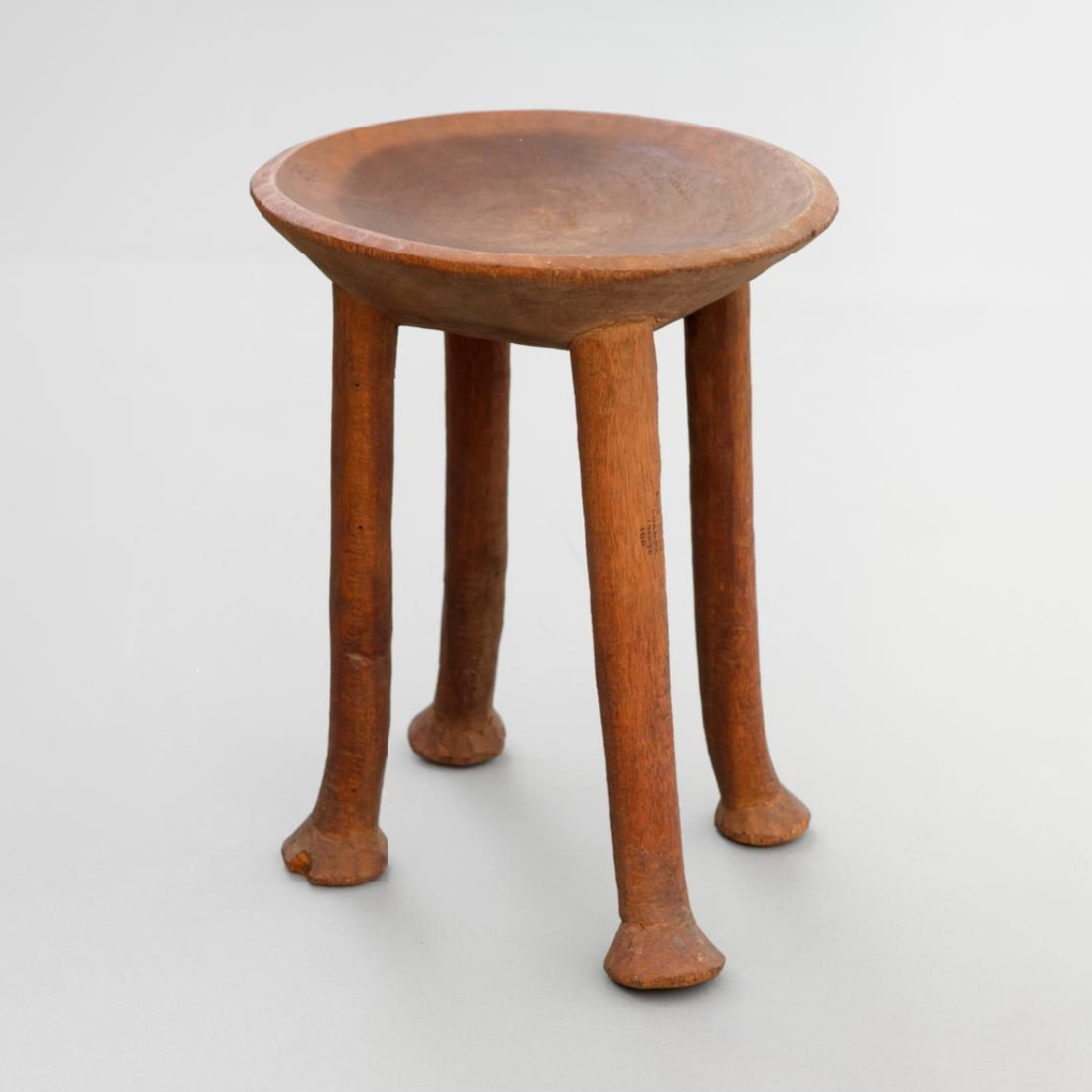The stool has four legs and a round dished top. Thew whole has been cut from a solid block of wood, each leg has a shallow conical foot.