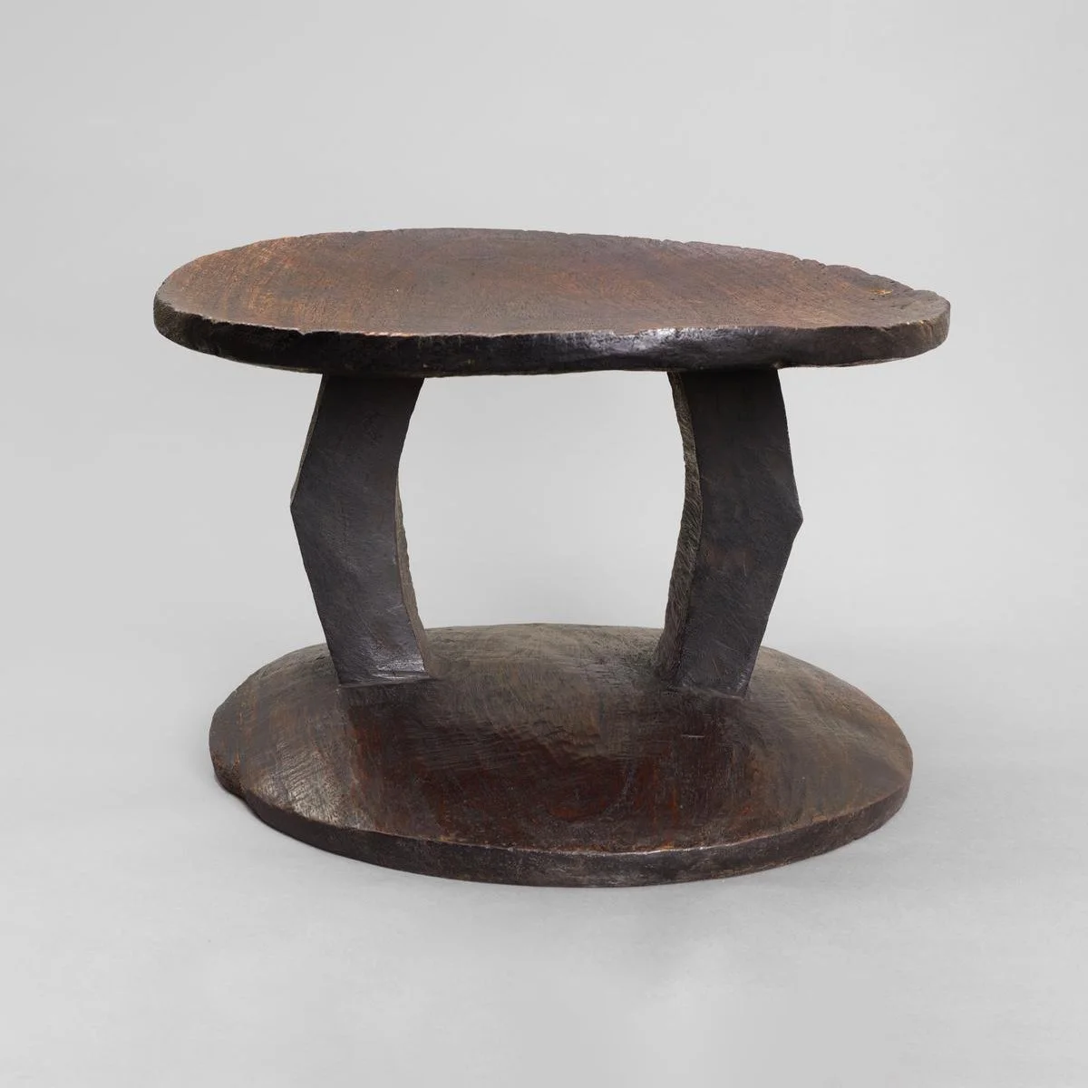 Stool with a top that is circular and dished, supported on two bent slightly angled square pillars. The base is round with a convex upper surface.