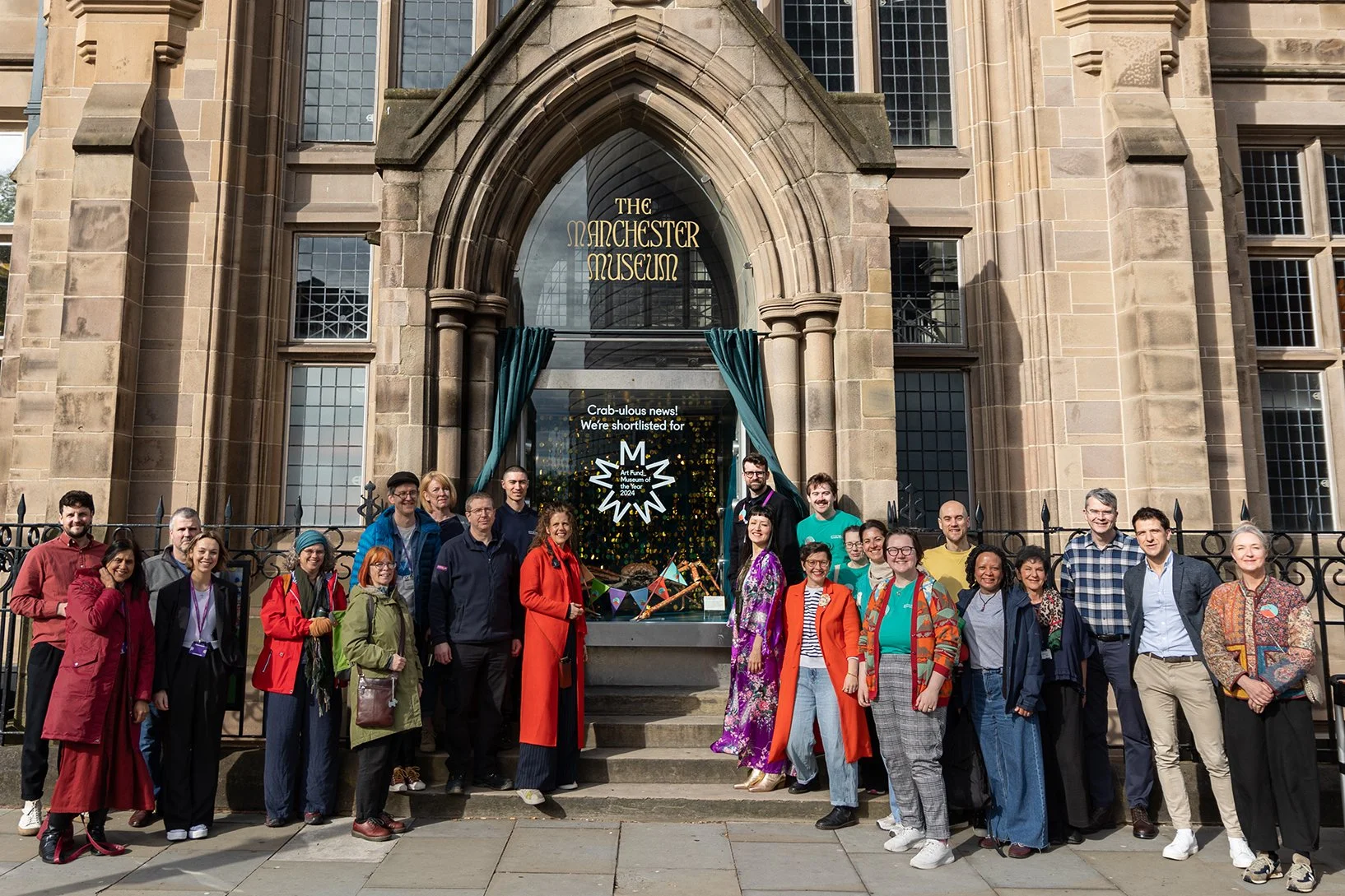 Group of people standing in front of the spider crab window outside Manchester Museum, in celebration of Manchester Museum's shortlisting for Art Fund Museum of the Year 2024.