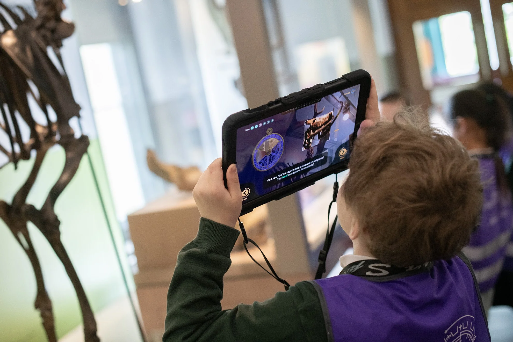 A young boy taking a photo or video with a tablet at a museum, with a skeleton of a large animal in the background.