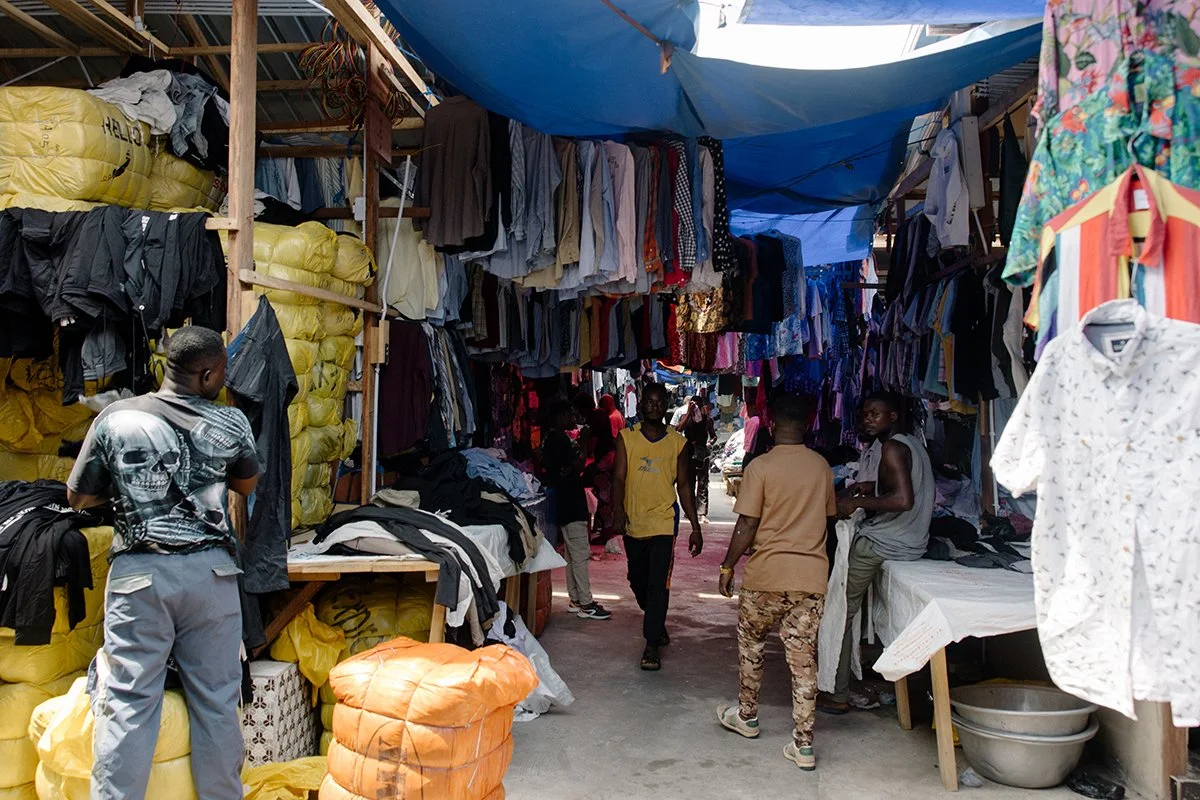 A busy clothes market showing people walking through stalls full of clothes, both hung and stacked in bales.