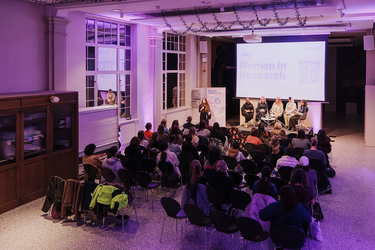 An audience listen to a talk on stage in the Main Hall at Manchester Museum. The space is lit in purple and there is a big screen behind the stage.