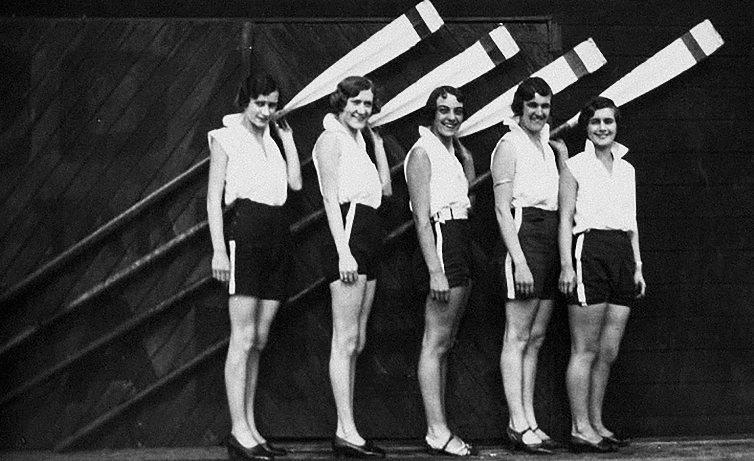 Grupo de mujeres deportistas sosteniendo remos, frente a una pared de madera, vestidas con ropa deportiva de época.