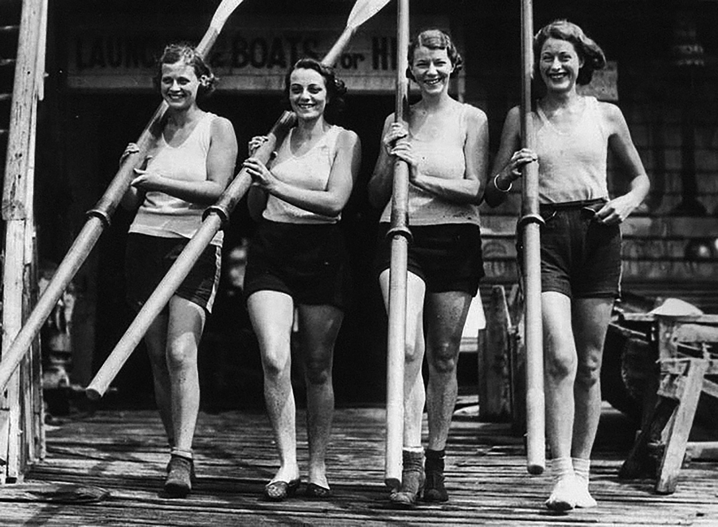 Cuatro mujeres jóvenes sonrientes sosteniendo remos, vestidas con camisetas sin mangas y pantalones cortos, caminando sobre un muelle de madera.