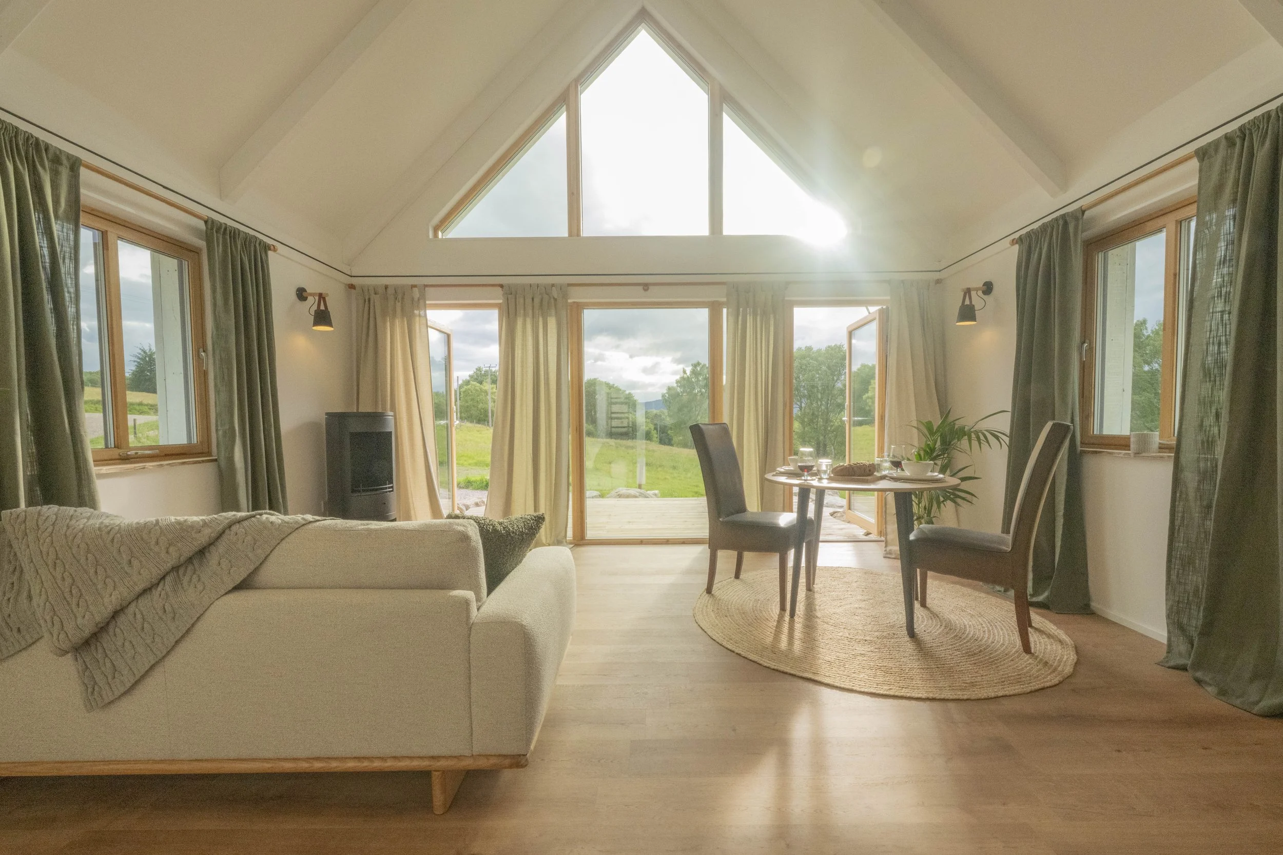 A spacious living room with large windows and glass doors, wooden floors, a beige sofa with a blanket, a round dining table with two chairs, a potted plant, and a view of greenery outside.