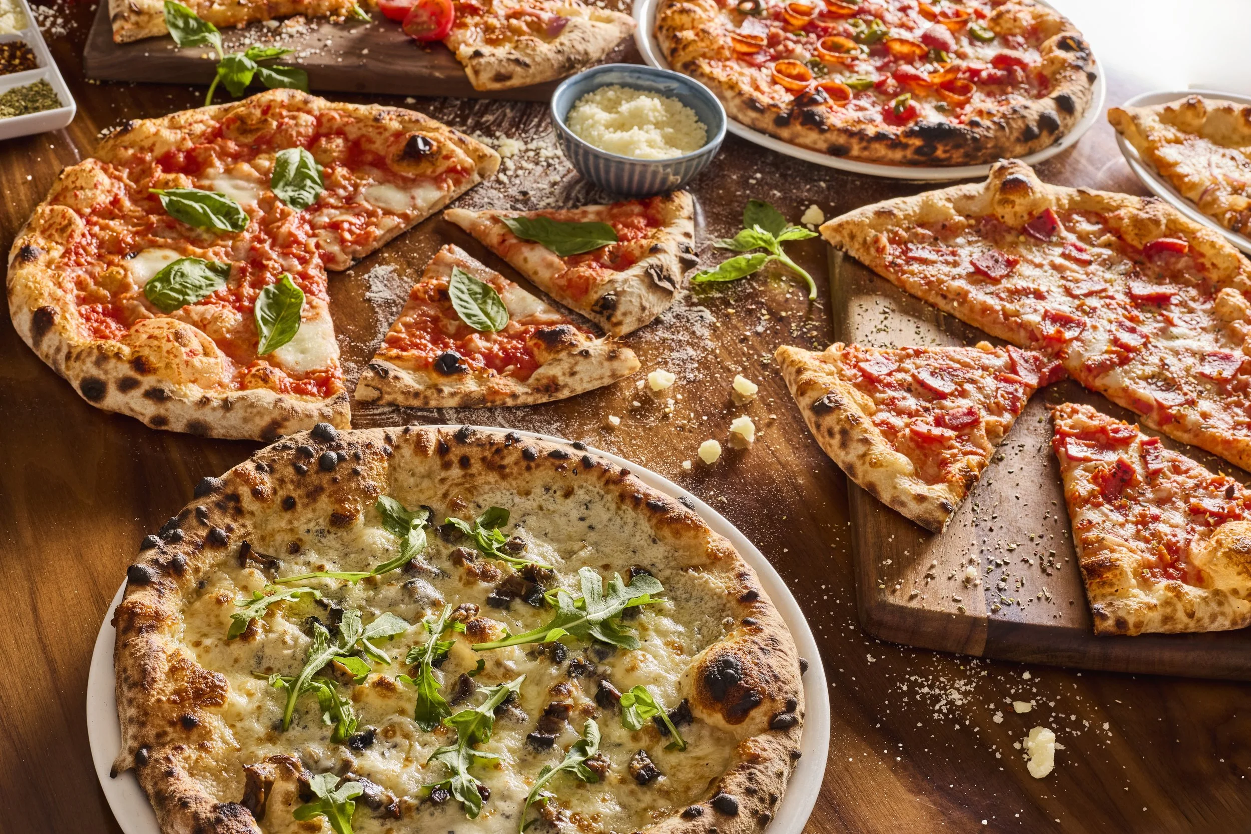 Various types of pizza on a wooden table, topped with cheese, herbs, and vegetables, with a bowl of grated cheese in the center.