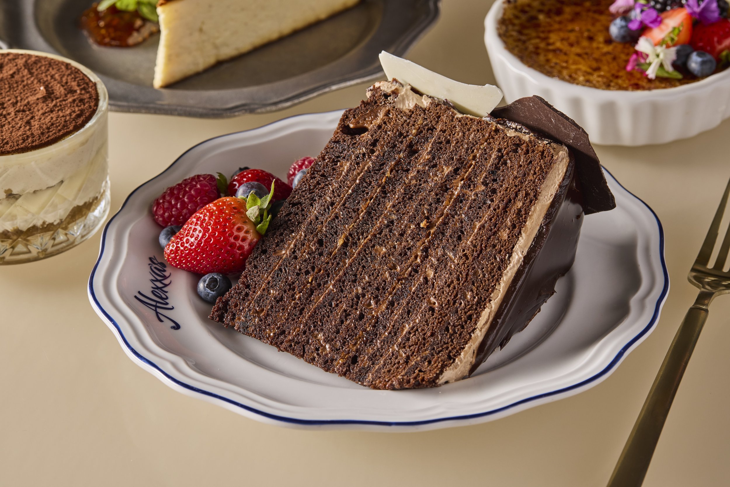 A slice of layered chocolate cake topped with a white chocolate shard, on a white porcelain plate with raspberry and blueberry garnishes, alongside a glass of coffee with cocoa powder and other desserts in the background.