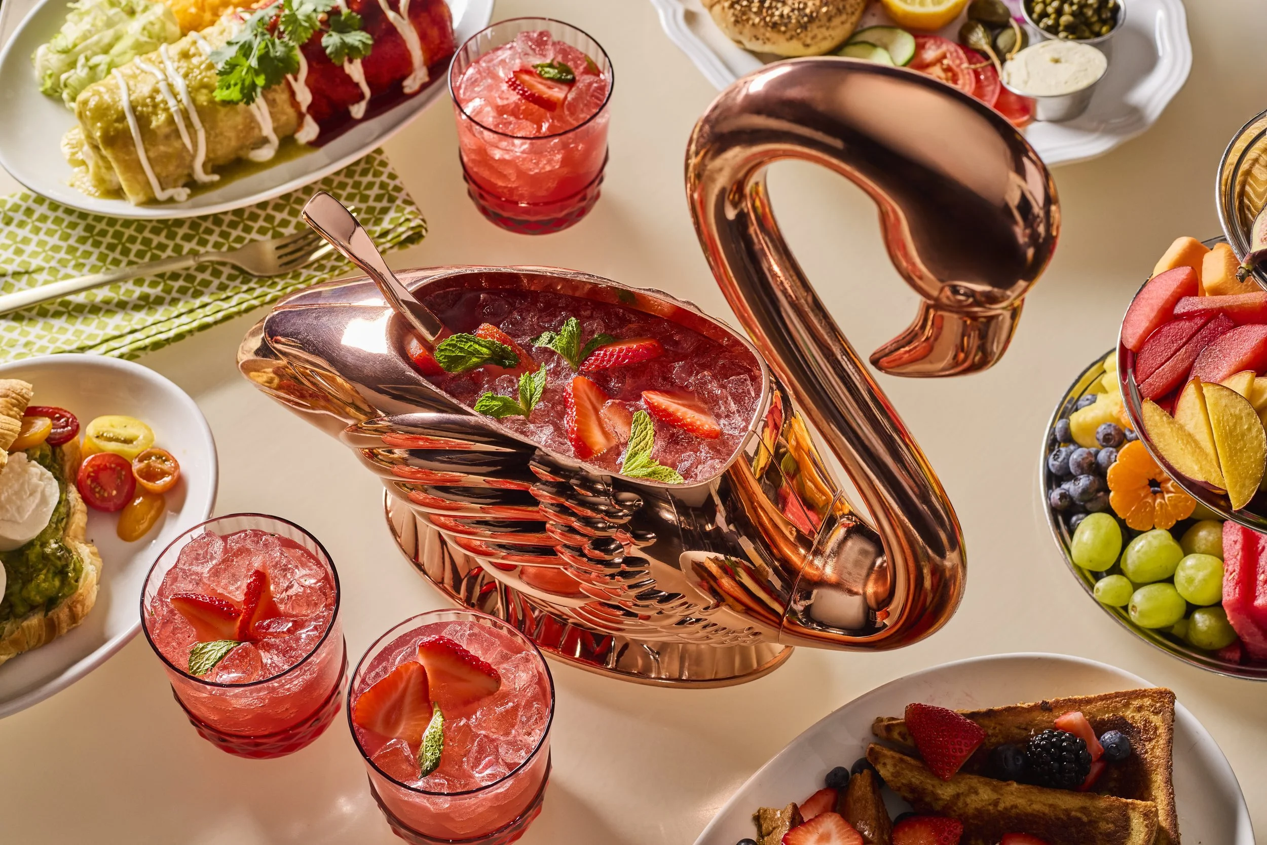An assortment of food and drinks including glasses of pink strawberry drinks, a fruit salad bowl with sliced fruits, a salad with cherry tomatoes, and a dessert with strawberries and mint leaves.