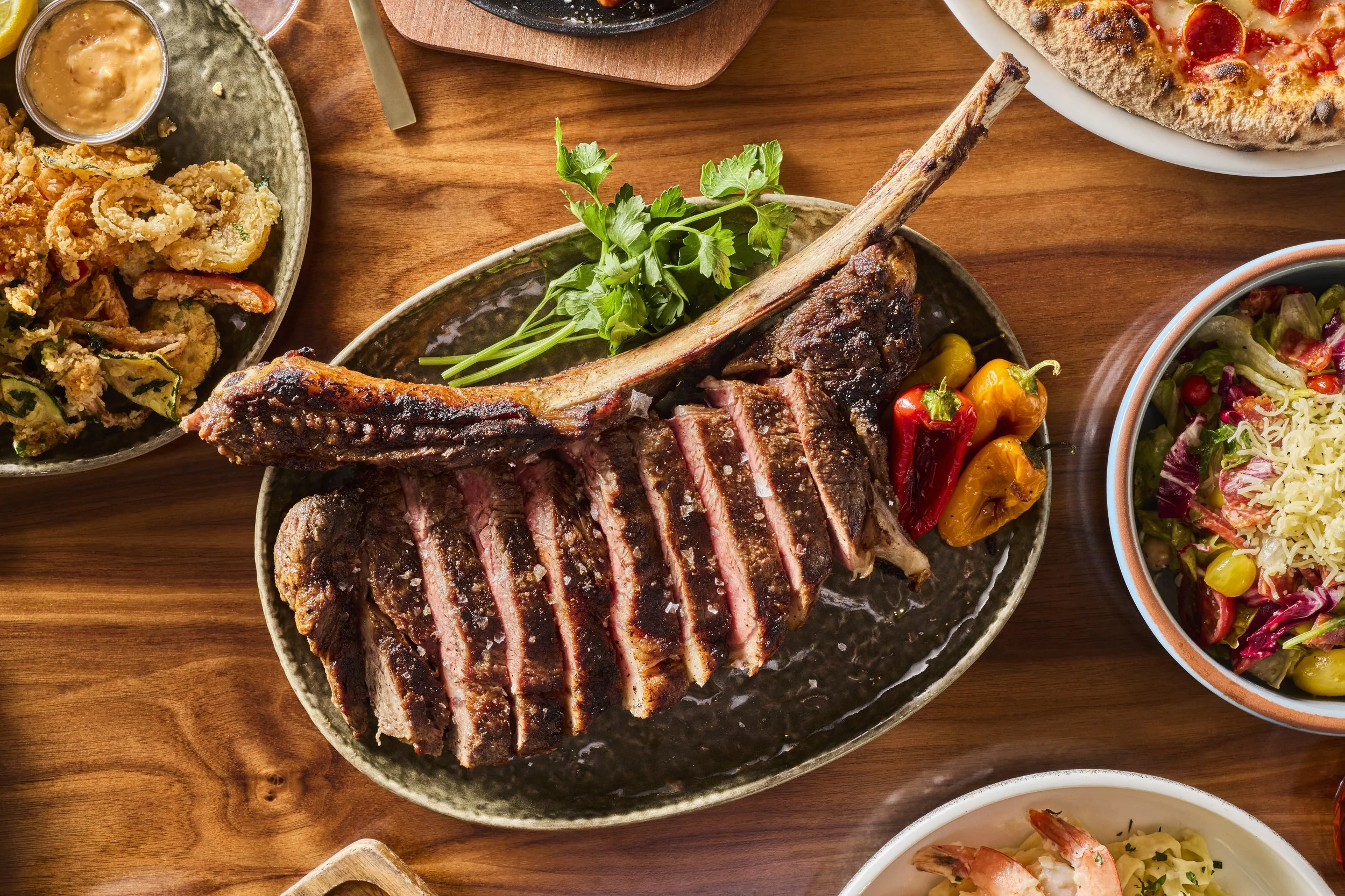 A spread of various dishes including grilled steak, fried calamari with dipping sauce, pizza, and salads served on a wooden table.