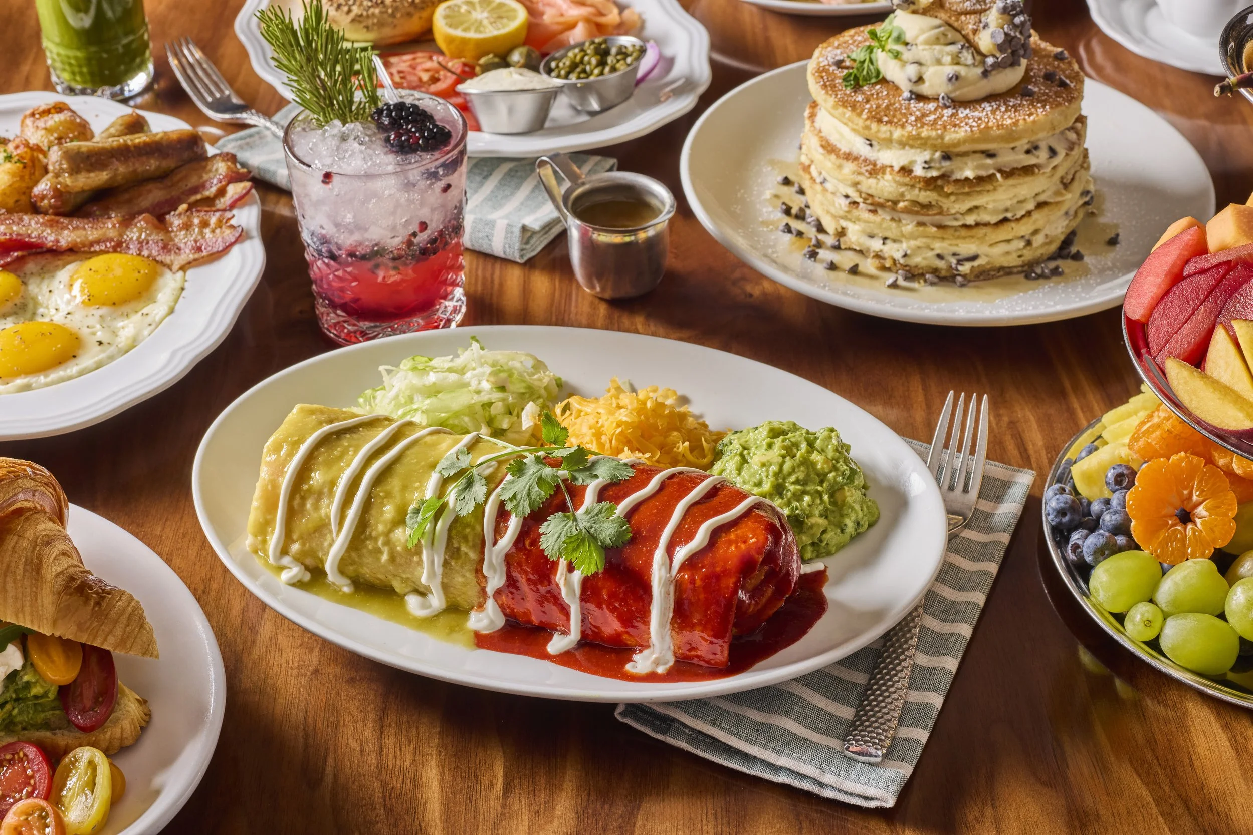 A table filled with various breakfast foods including pancakes with chocolate chips, a plate of eggs, bacon, sausages, and vegetables, a fruit bowl with grapes and orange slices, and a colorful drink with berries and rosemary.