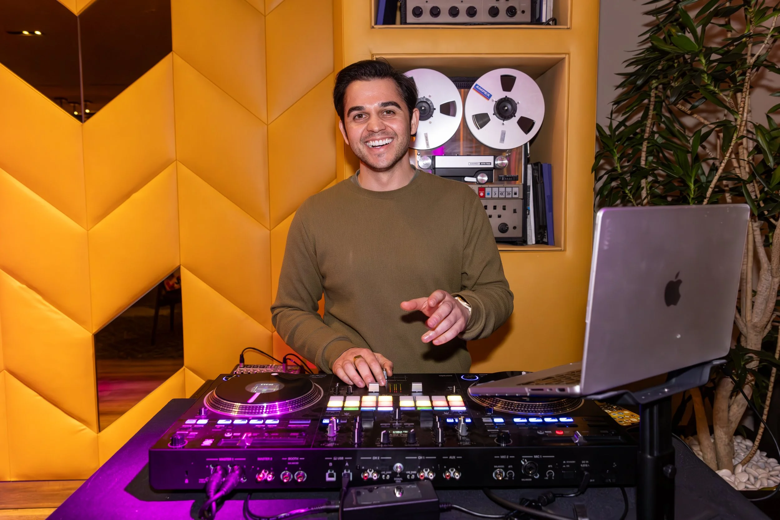 A man smiling and DJing with a DJ controller, laptop, and speakers behind him, in a room with yellow padded wall panels and a large leafy plant.
