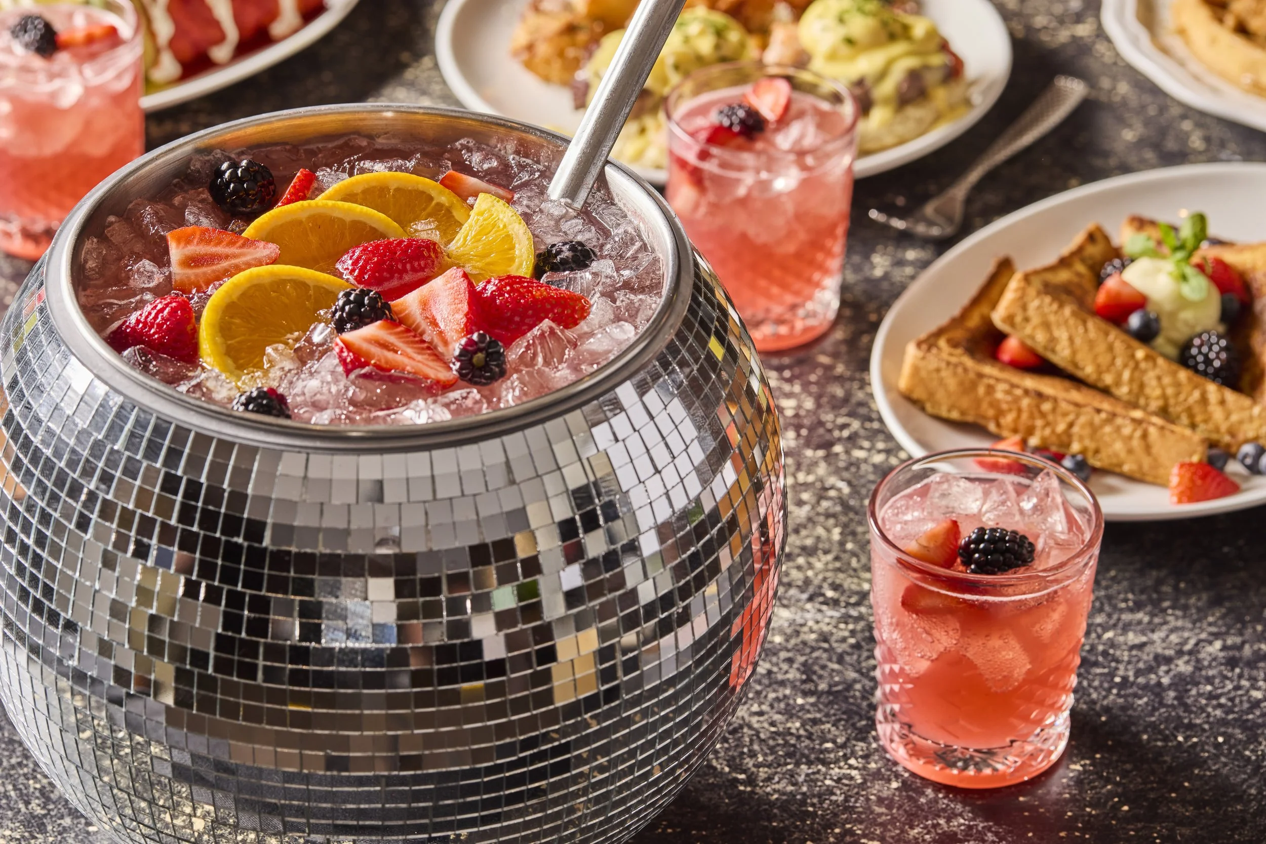 A beverage dispenser filled with pink drink topped with orange slices, strawberries, and blackberries. Surrounding plates of food including French toast with berries and whipped cream, pasta with sauce, and a pink cocktail garnished with fruit are on a dark, speckled table.