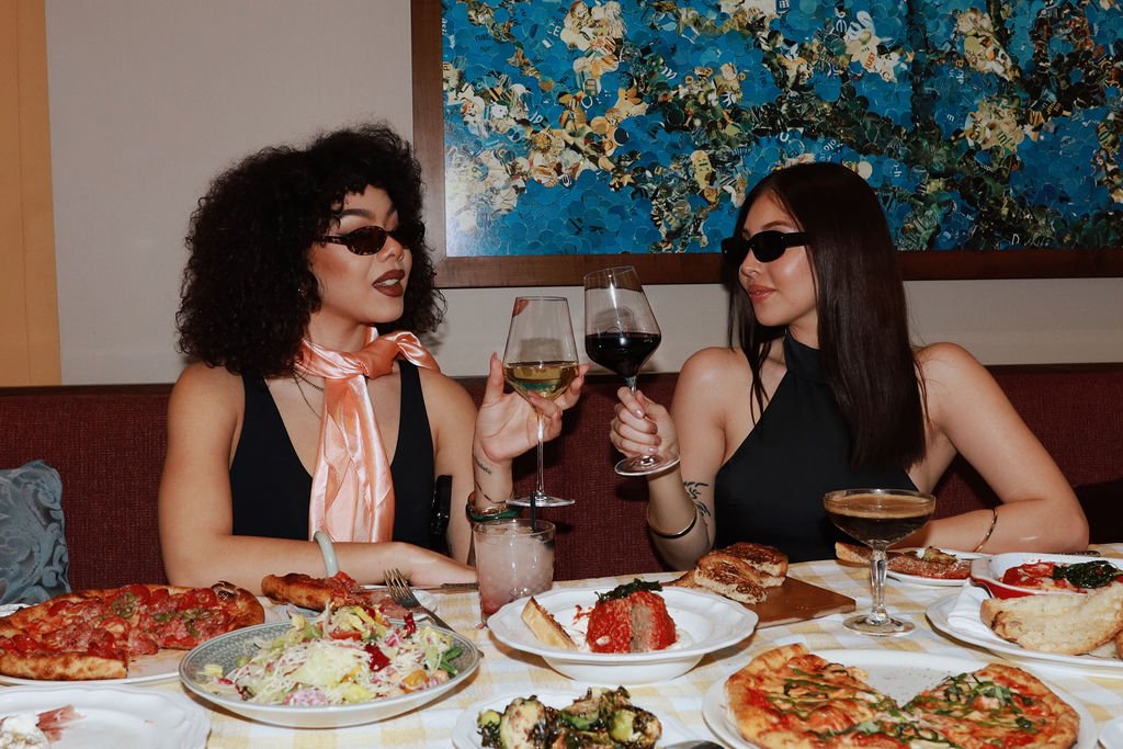 Two women wearing sunglasses enjoying wine and pizza at a restaurant table with various dishes and a colorful abstract painting in the background.