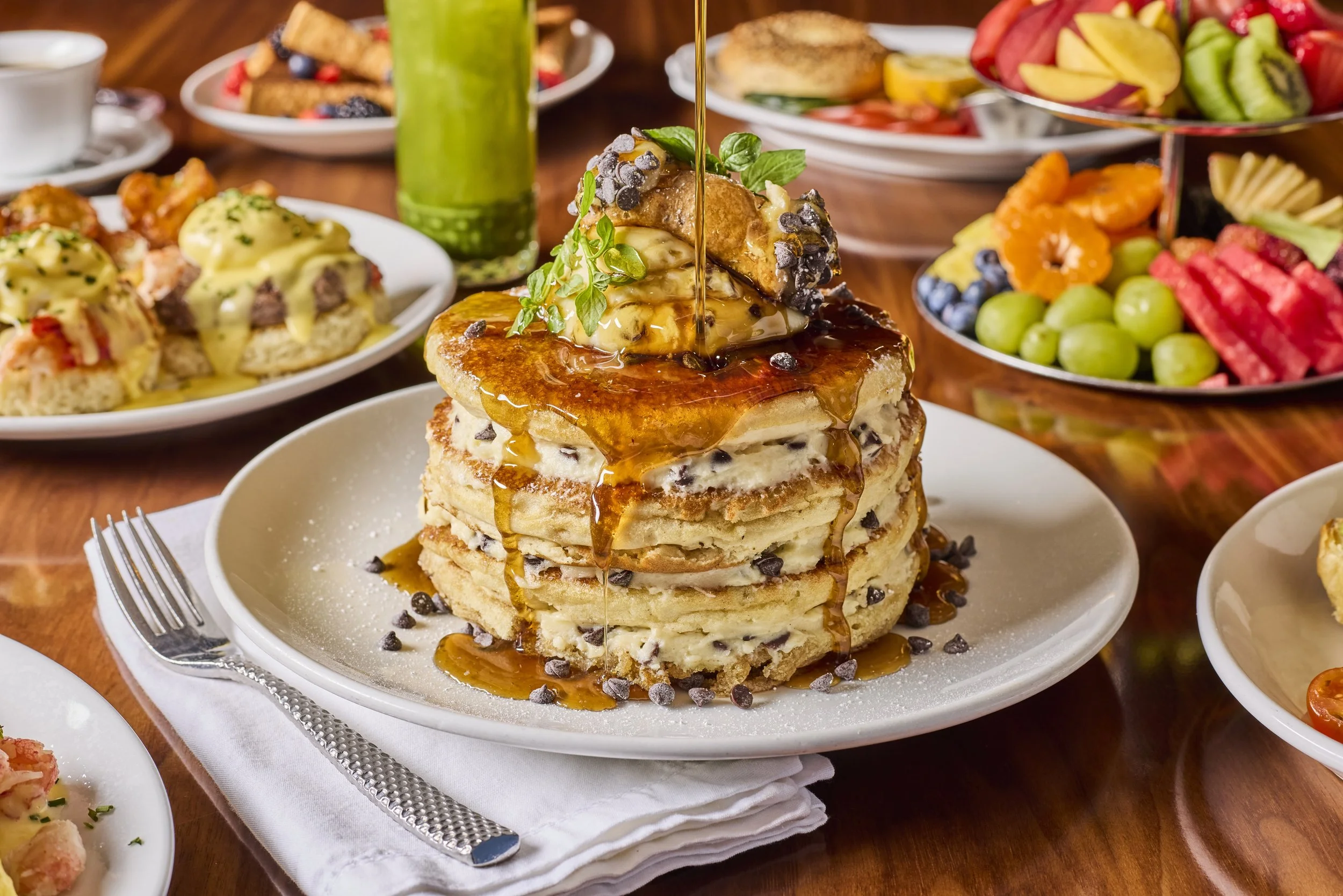Stack of chocolate chip pancakes topped with syrup, whipped cream, chocolate chips, and fresh mint, surrounded by plates of fruit, breakfast dishes, and a green smoothie on a wooden table.