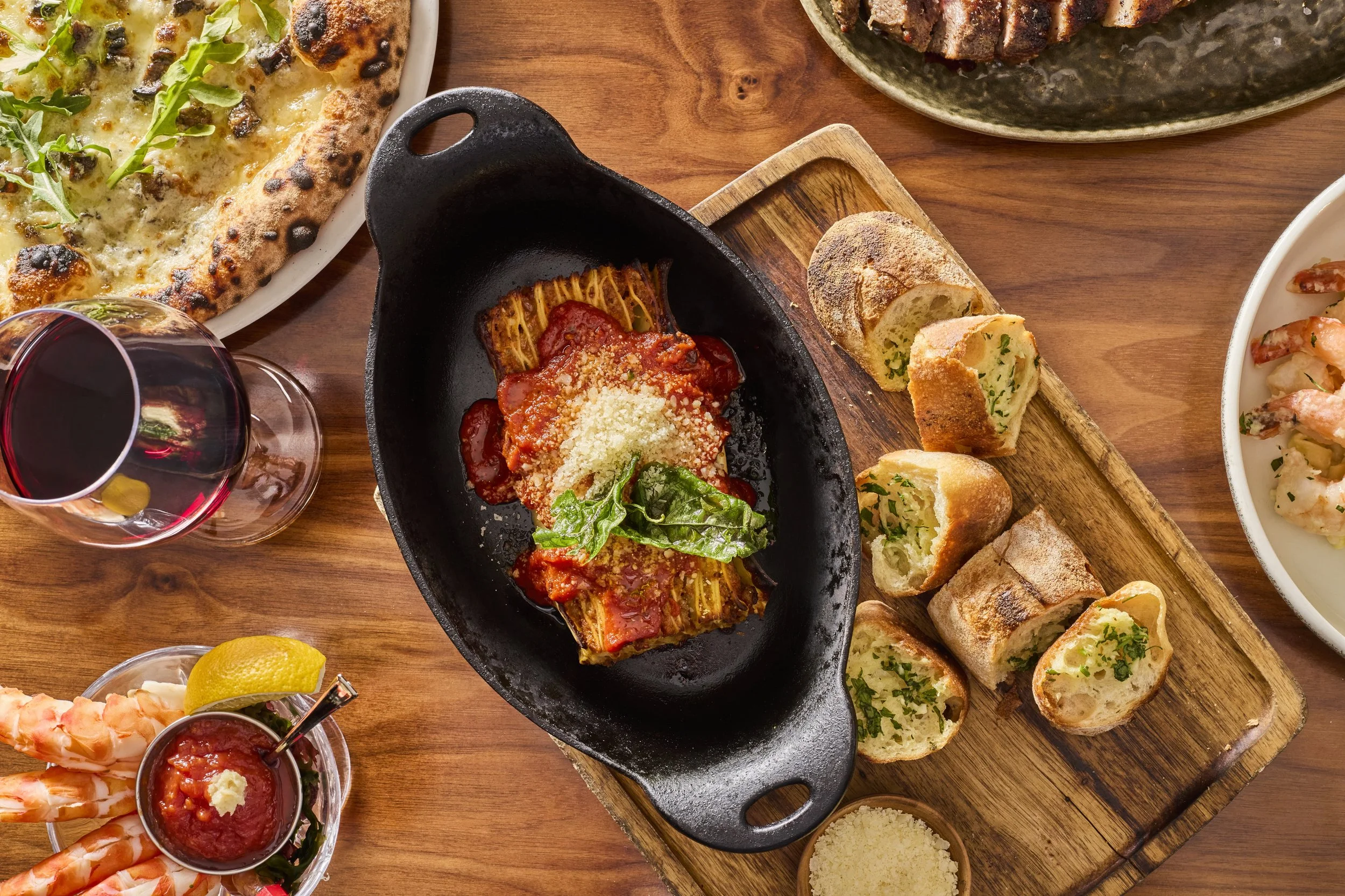Various Italian dishes on a wooden table, including pizza, pasta, garlic bread, shrimp, and a glass of red wine.