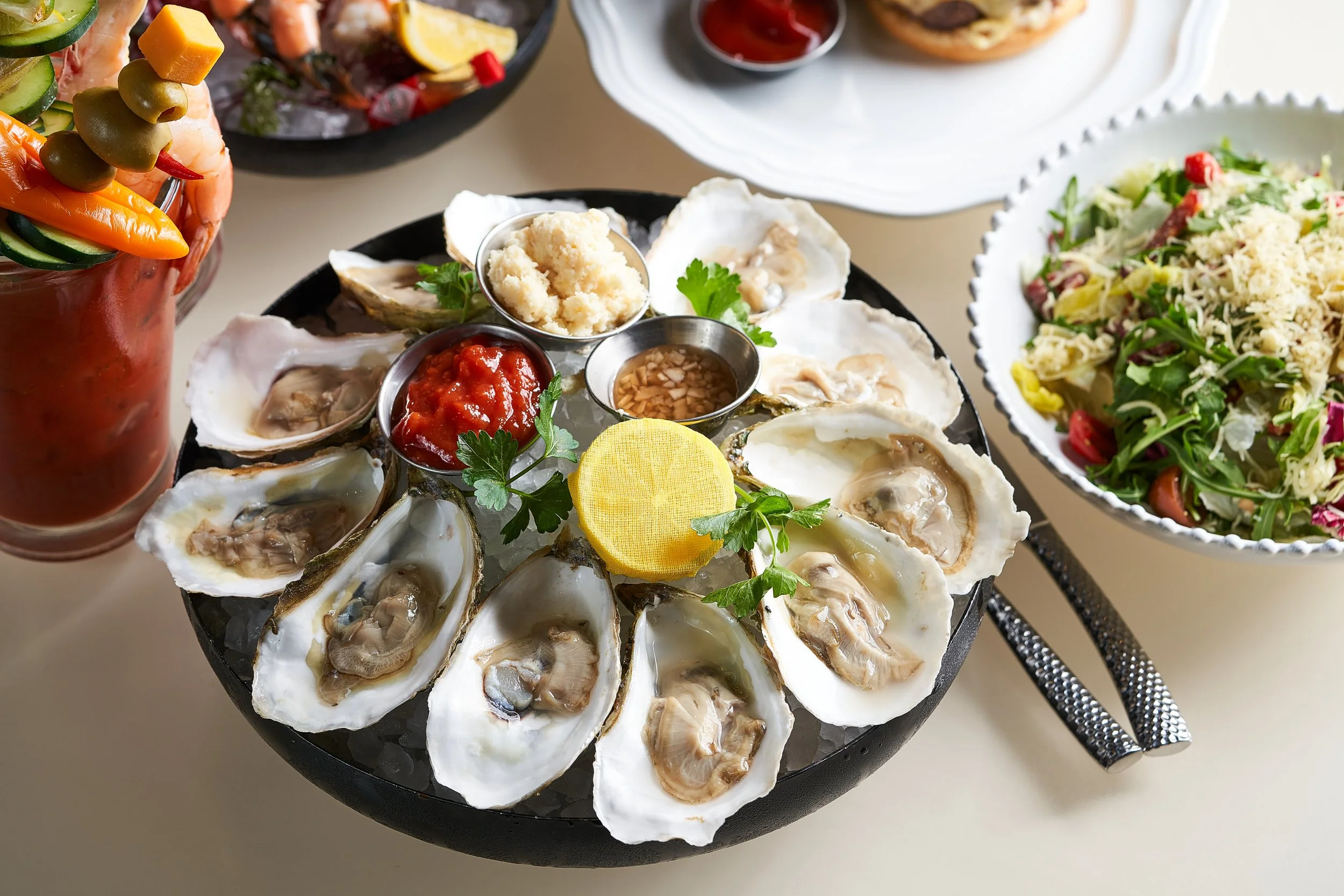 A black platter with fresh oysters on ice, garnished with lemon slices, parsley, red sauce, and various condiments, surrounded by side dishes including salad, complementary sauces, and a jar of vegetable sticks.