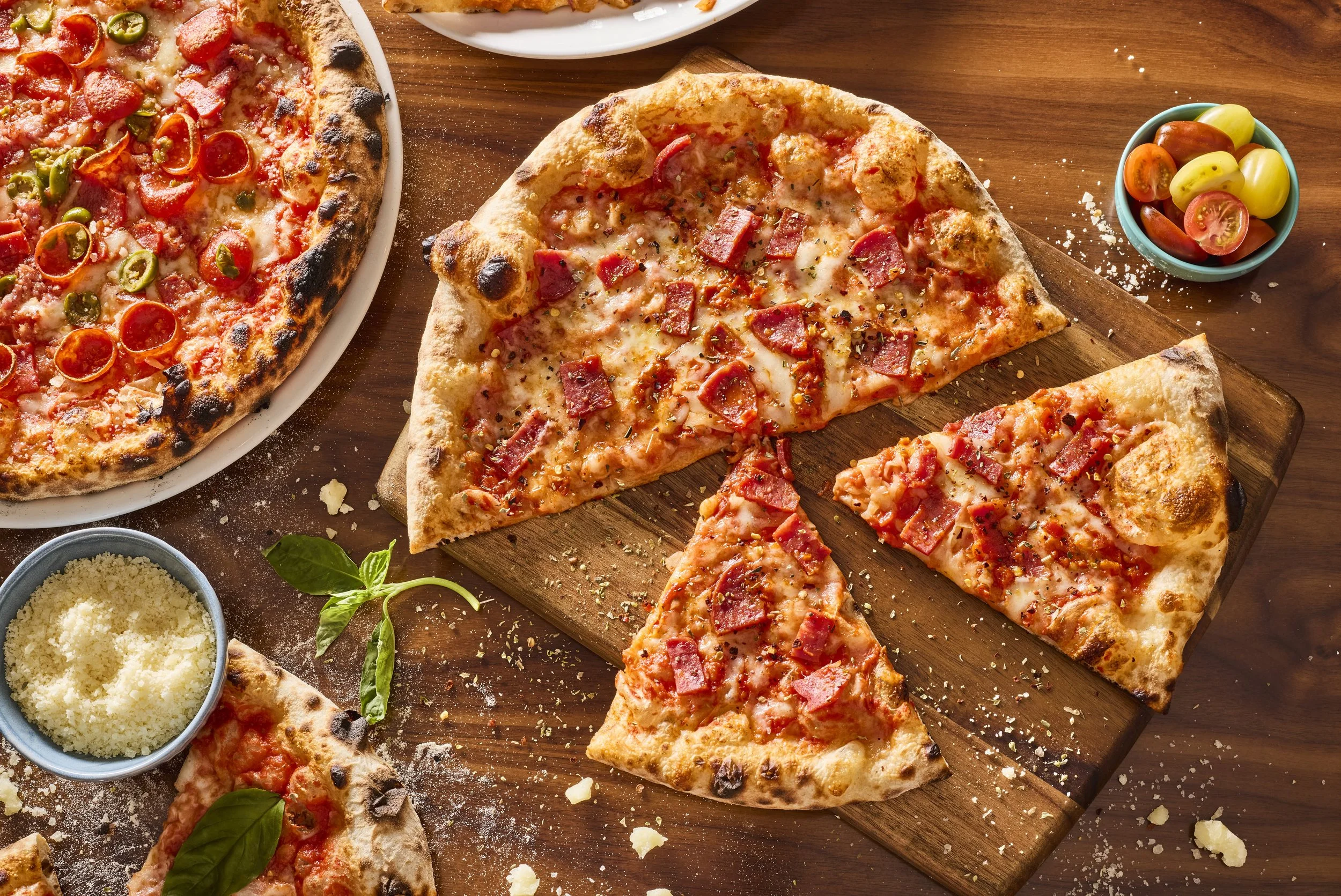 Several slices of pepperoni and ham pizza on a wooden cutting board, with a side bowl of cherry tomatoes and grated cheese around.