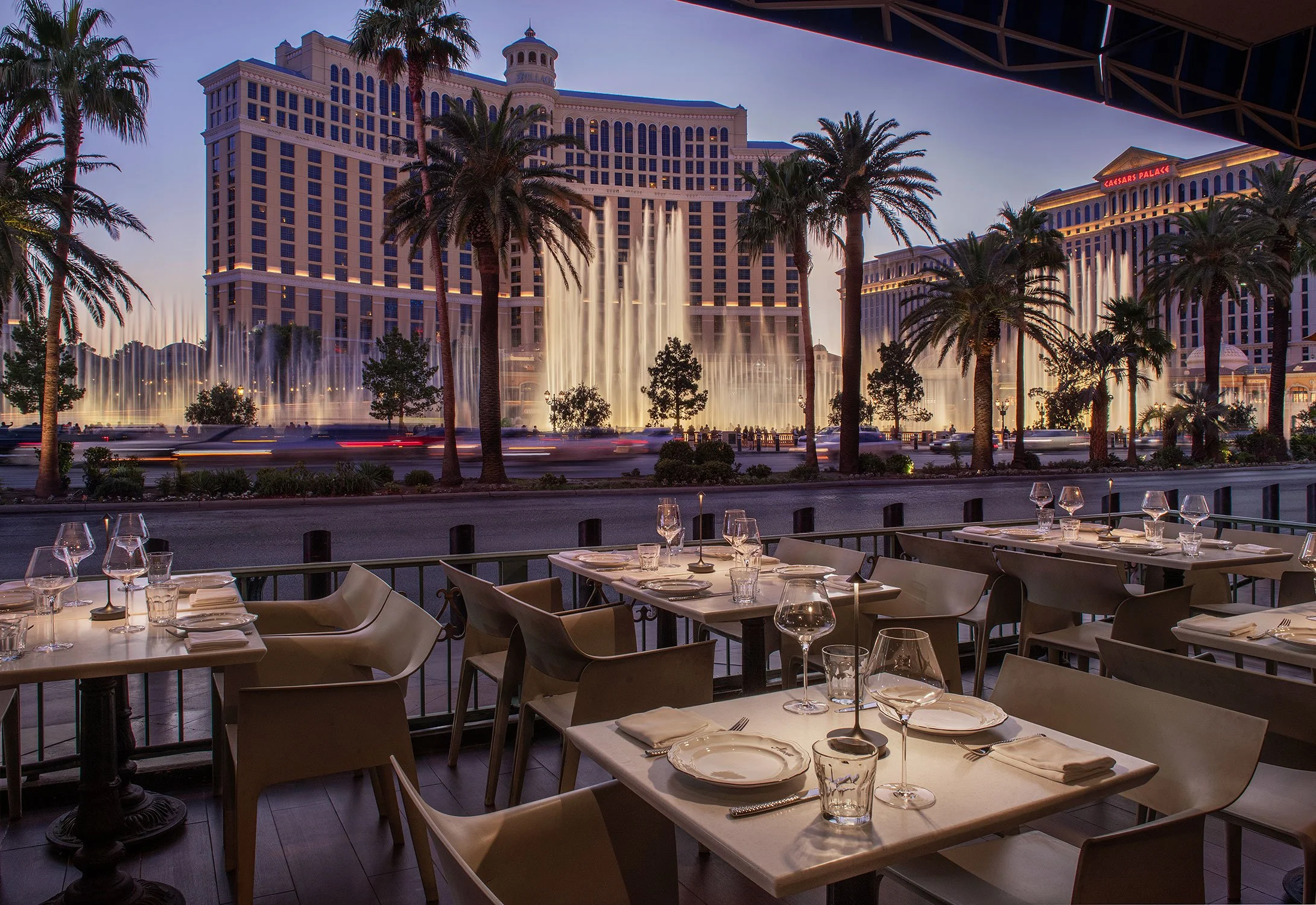 Outdoor dining area at a restaurant overlooking the Bellagio hotel and fountains in Las Vegas, with tables set with glasses and napkins, during evening with the hotel lit up and fountain display in progress.