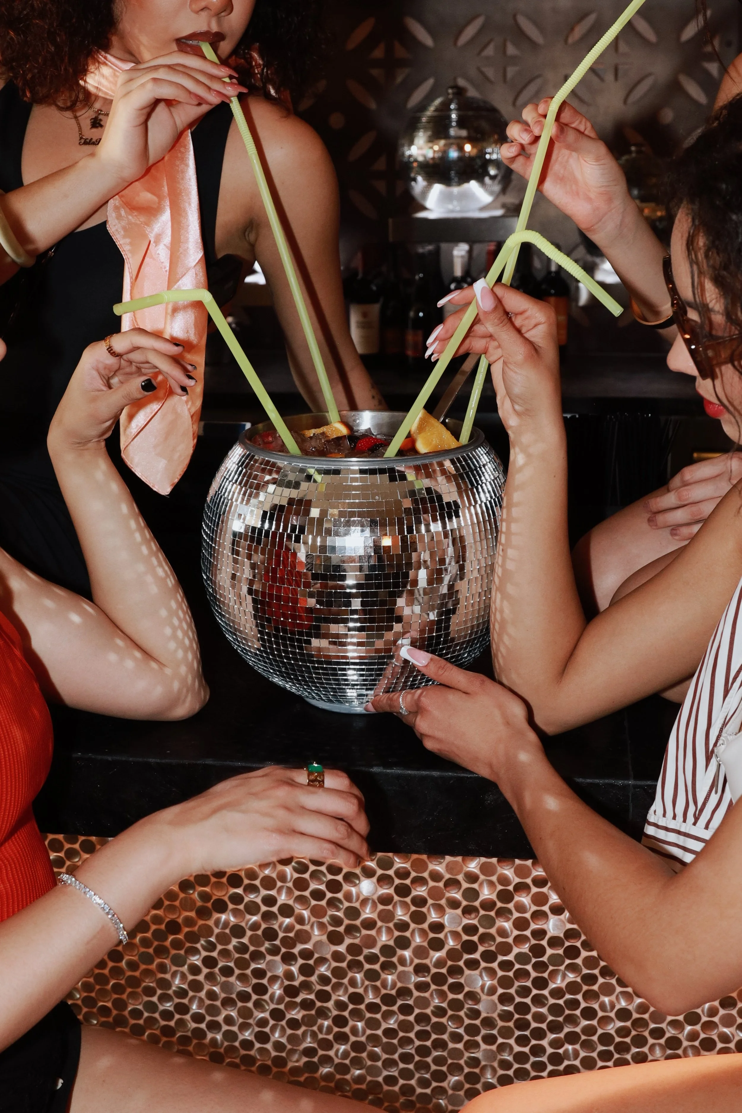 Four women are gathered around a shiny, disco ball-style container filled with drinks and fruit, using large, colorful bendy straws to share the beverage.